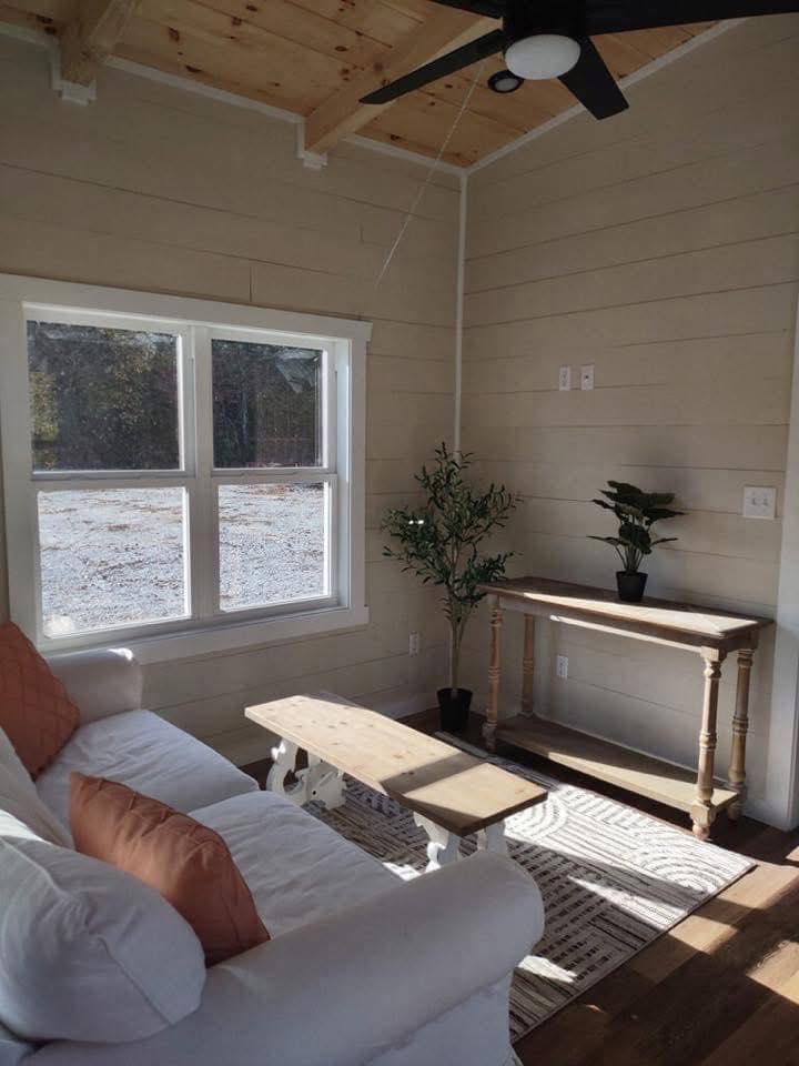 Cozy living room with a white couch and orange cushions, wooden coffee table, and a console table with plants. Sunlight filters through a large window.