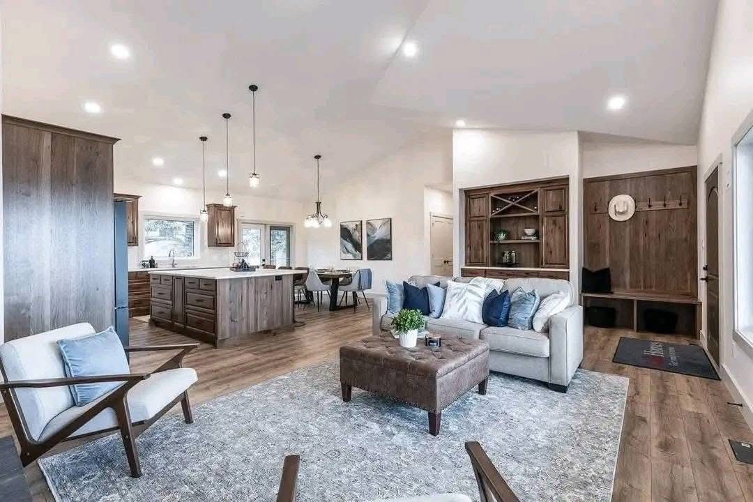 Spacious modern living room with stylish wood accents. A cozy sofa with blue pillows faces a central ottoman. Bright, inviting, with sleek kitchen area.