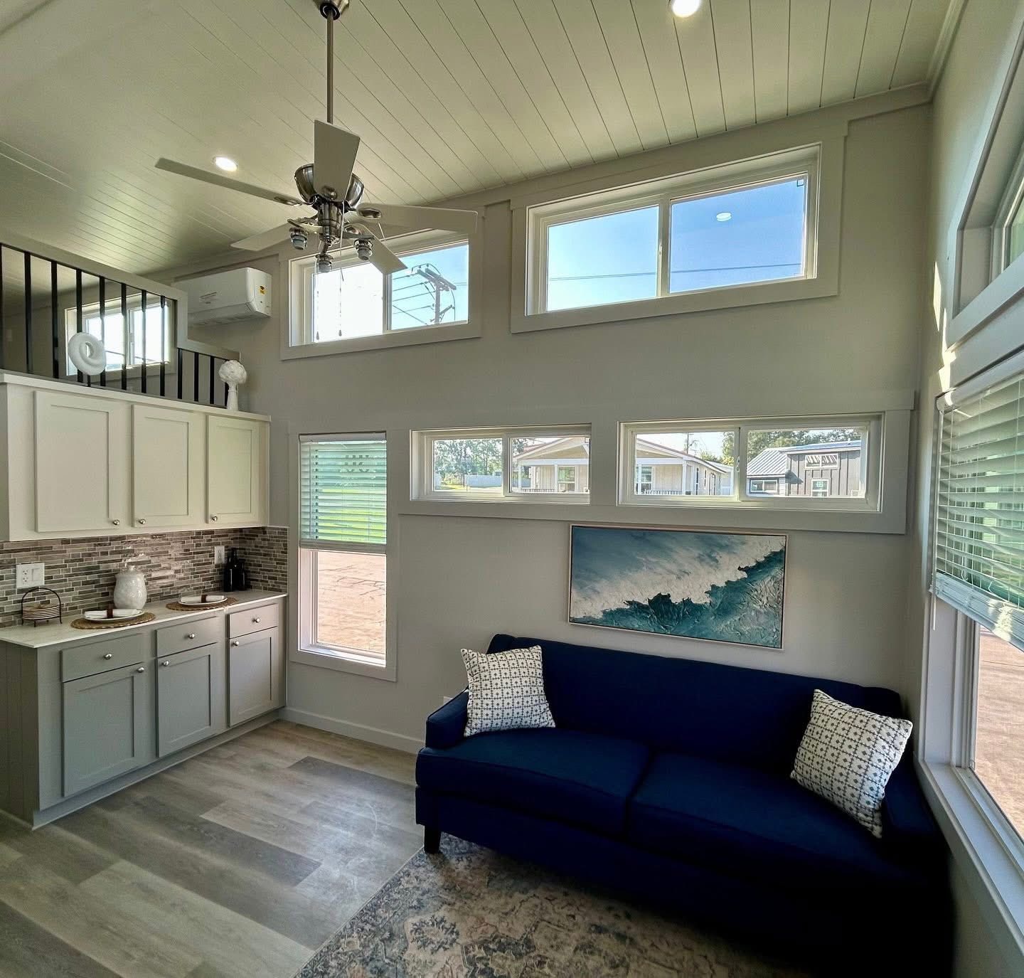 Bright, modern living space with high ceilings, lots of windows, and a blue sofa. Kitchenette with white cabinets, sleek decor, and a tranquil vibe.