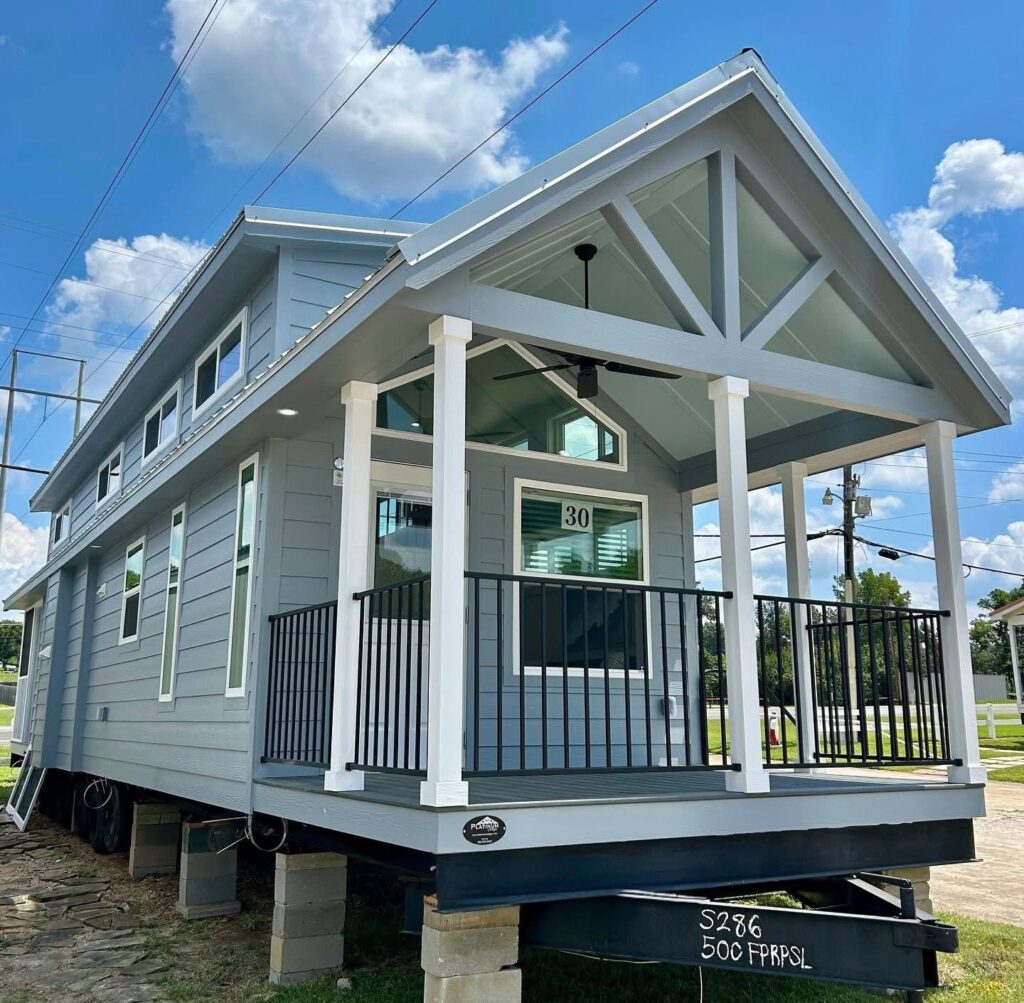 A modern grey tiny house on raised blocks with a covered porch featuring a gable roof, black railings, and white trim. Blue sky and clouds in the background.