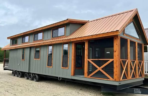 Modern tiny house on wheels with green siding and a warm, wood-framed porch. It has multiple windows and a metal roof, set in a gravel area under a cloudy sky.