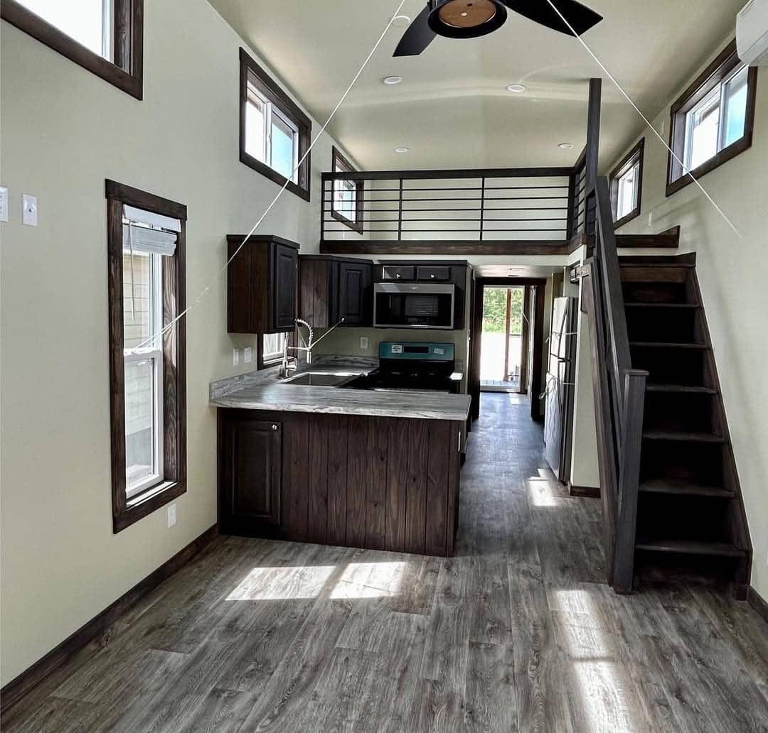 Modern tiny house interior with a loft. Features dark wood accents, stairs, a kitchen with an island, and multiple windows, creating an open, airy feel.