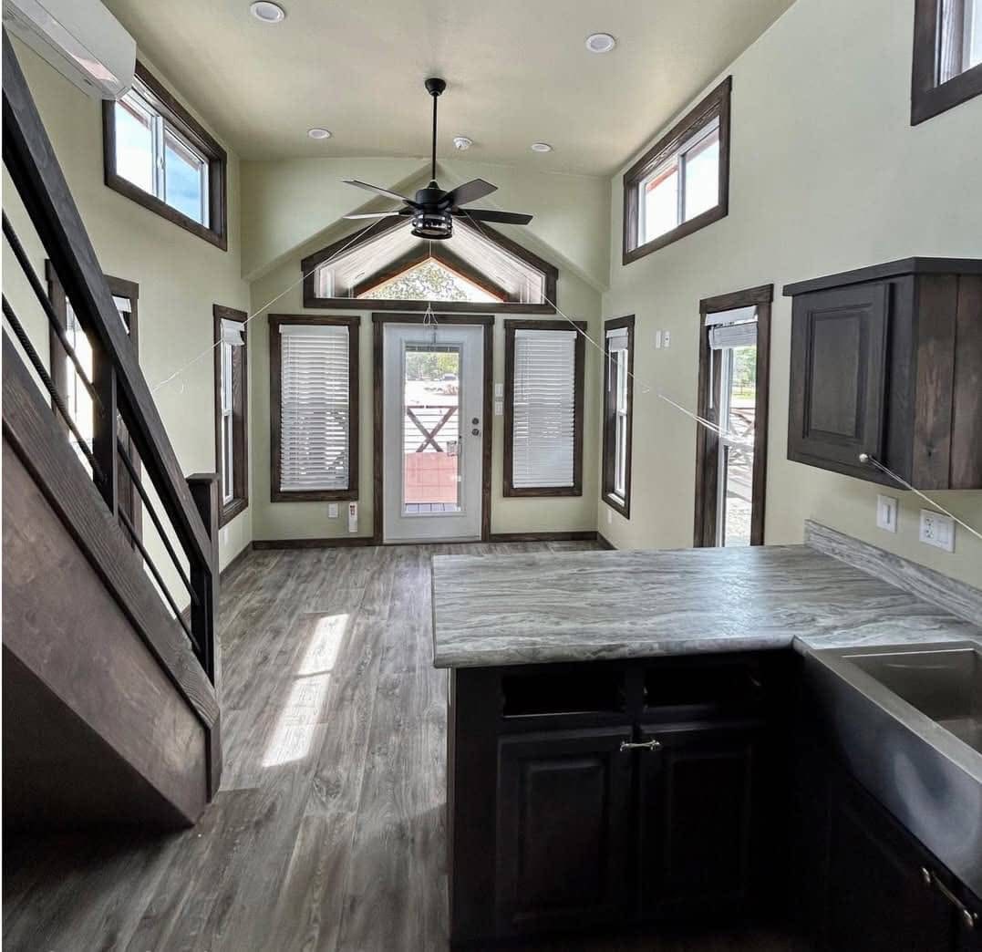Interior of a modern tiny home with high ceilings. Features light-filled space with large windows, a ceiling fan, and dark wood accents. Cozy and open.