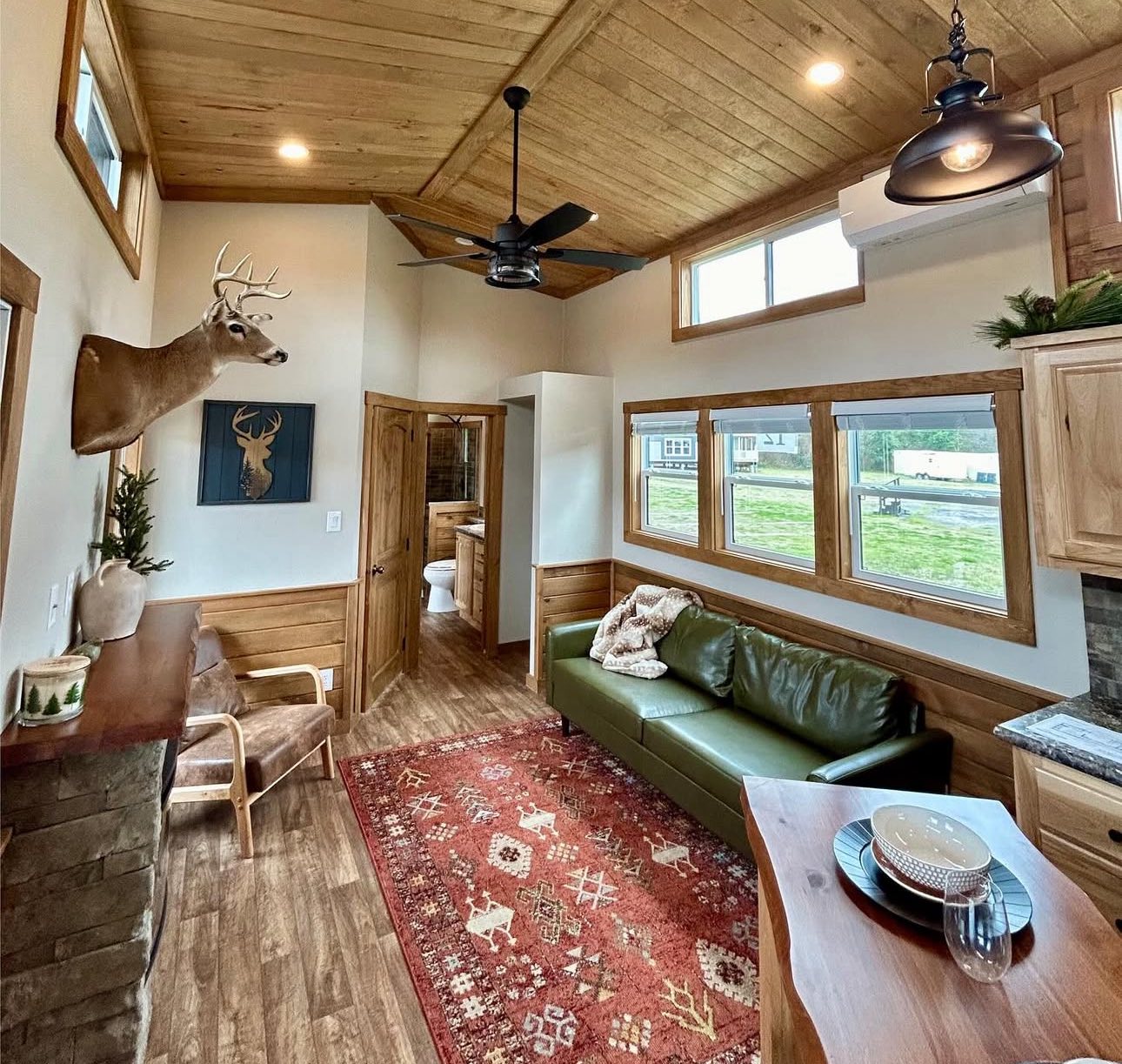 Cozy rustic living room with a vaulted wooden ceiling, green couch, red patterned rug, deer mount on the wall, and large windows overlooking greenery.