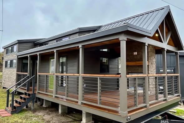 Modern tiny house with a sleek design features dark wood siding, metal roof, and a spacious covered porch with railings, set in a grassy area.