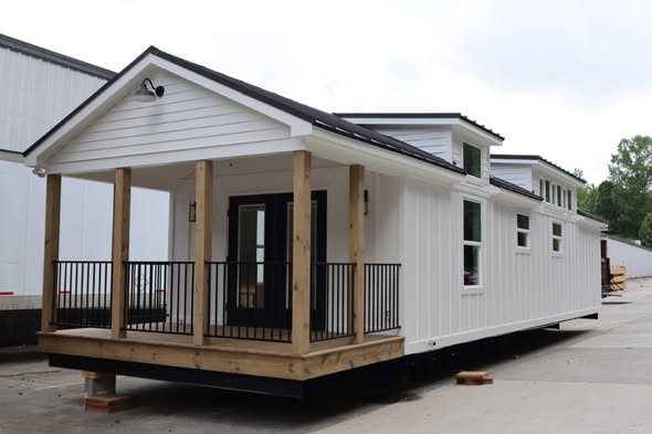 White, modern tiny house on a lot, featuring a gabled roof, wooden porch with black railings, and multiple windows. The scene is calm and tidy.