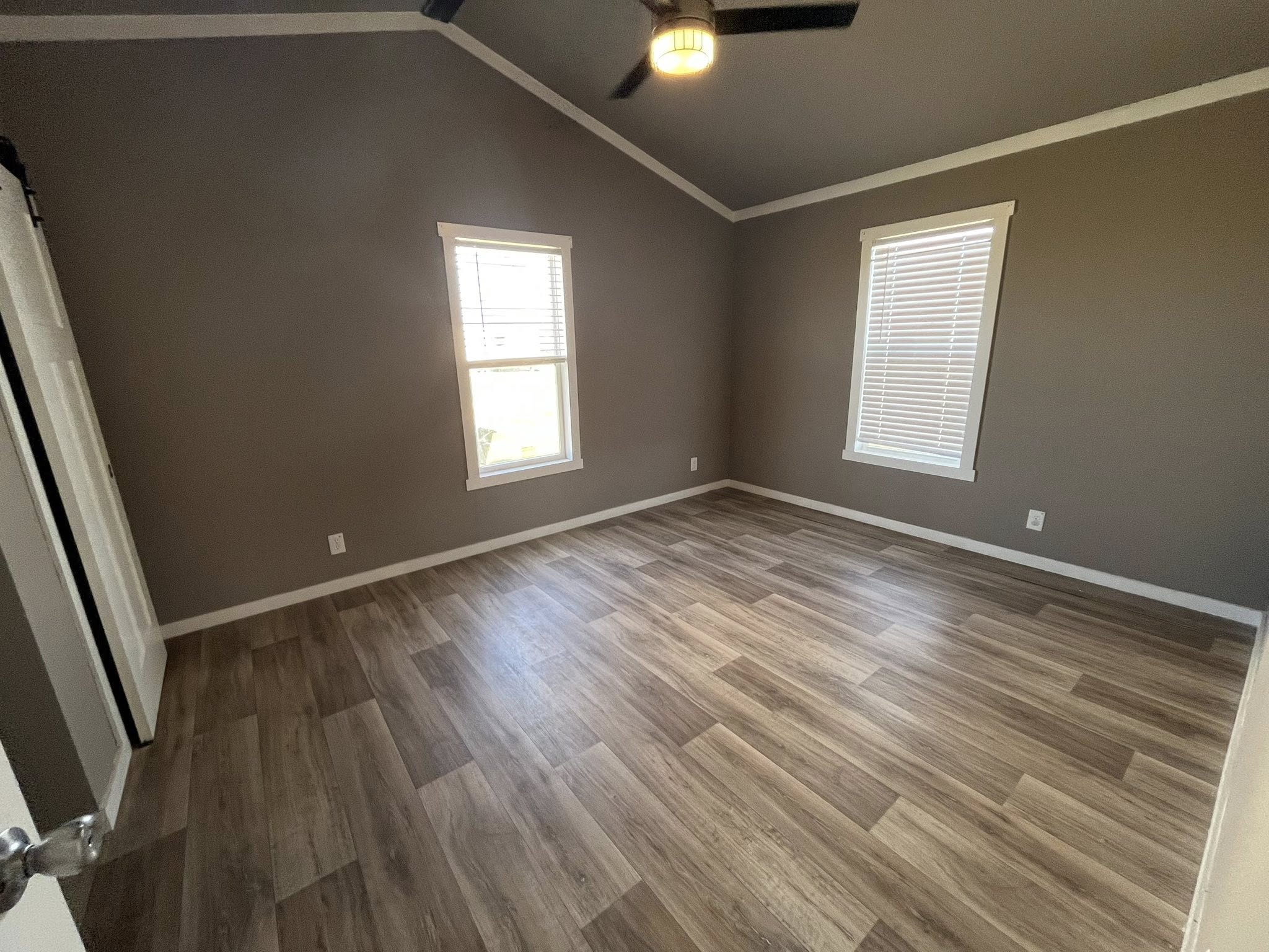 Empty room with light wood flooring and two windows with white blinds against taupe walls. A ceiling fan with a light is visible, creating a cozy atmosphere.