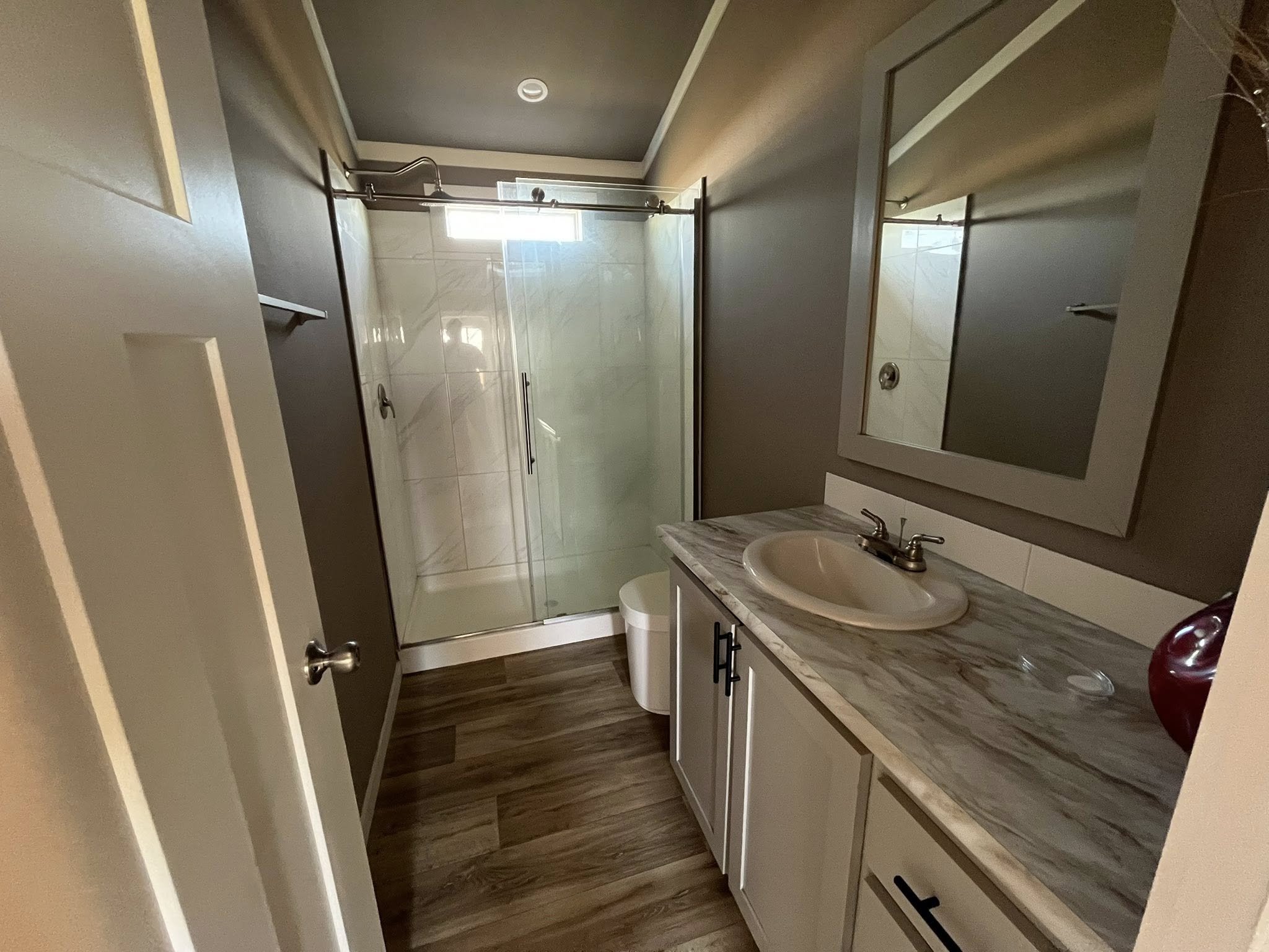 Narrow bathroom with wooden floor, a marble countertop sink on the right, a large mirror, and a glass-enclosed shower at the back. Warm, cozy feel.