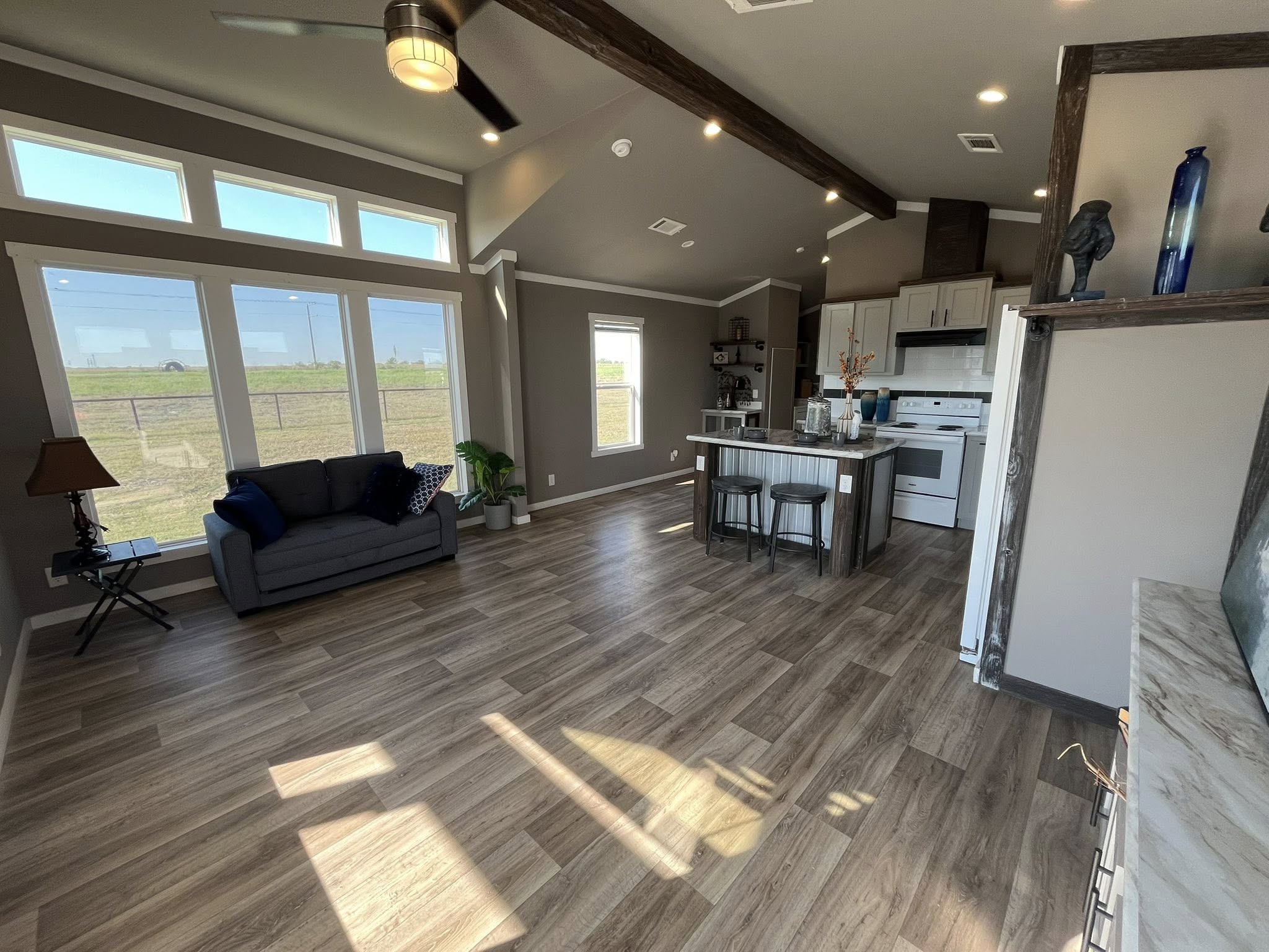 Spacious living area with large windows, wood flooring, a cozy gray sofa, and a modern kitchen. The room feels bright and inviting.