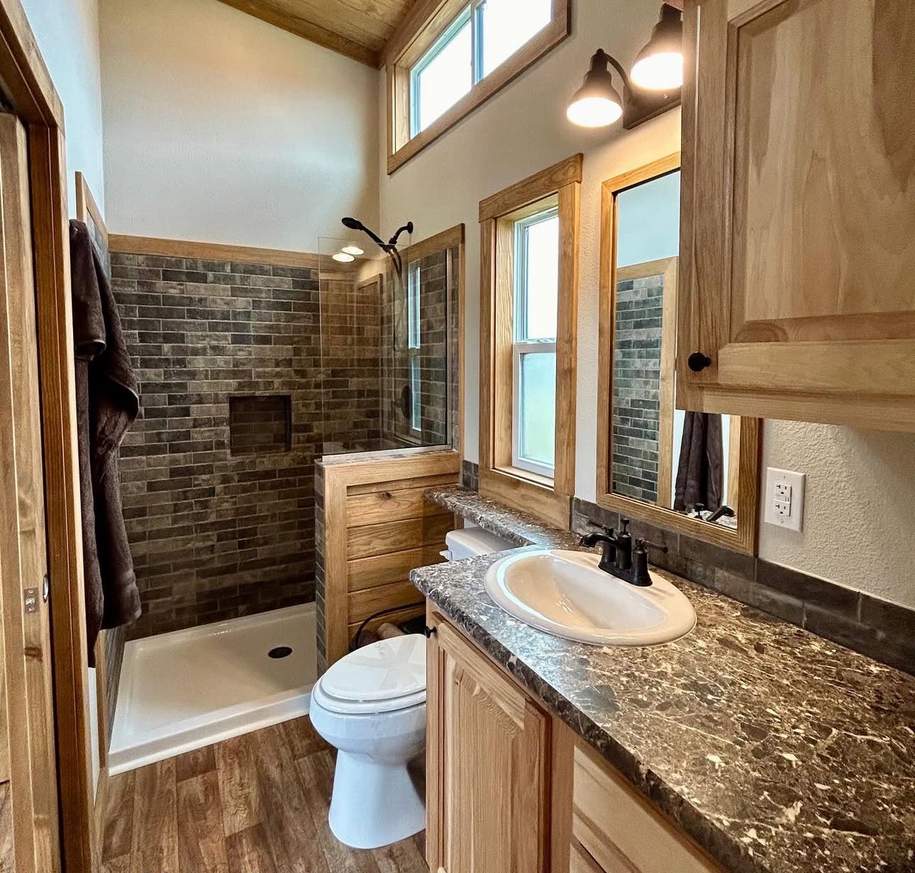 Compact bathroom with rustic wood accents, featuring a marble countertop sink, wood cabinets, brick-tiled shower, and wood flooring. Warm ambiance.