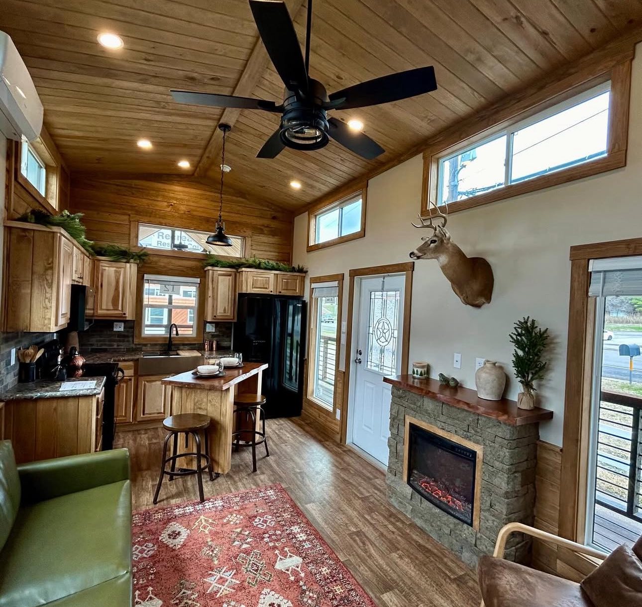 Cozy tiny home interior with wooden ceiling and floors. Features a kitchen with wooden cabinets, black fridge, and island. A mounted deer head is above an electric fireplace, and a green sofa faces a patterned rug, creating a warm, rustic ambiance.