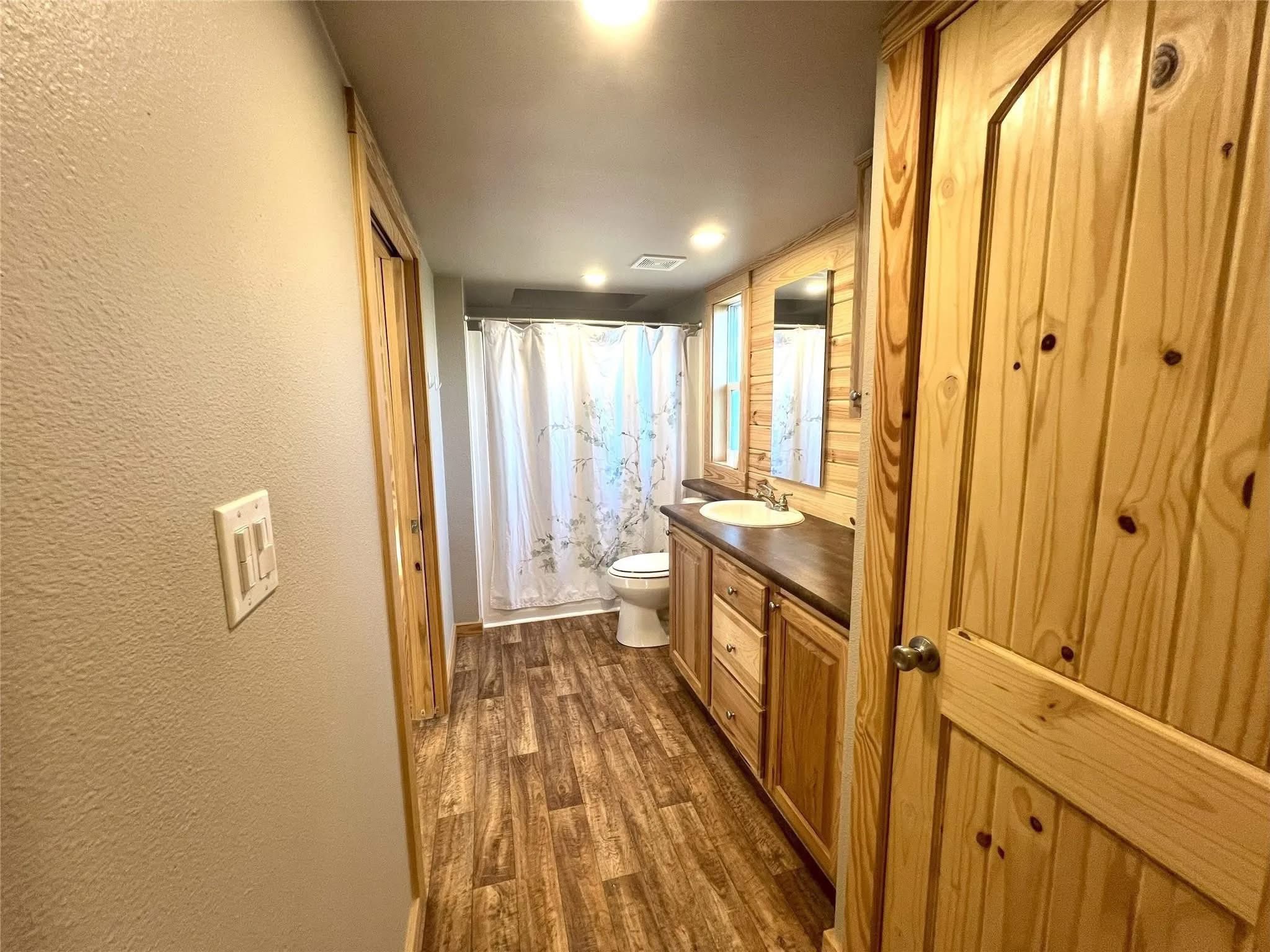Cozy bathroom with wood paneling, featuring a wooden door, floor, and cabinets. A shower with a floral curtain and sink add to the inviting feel.