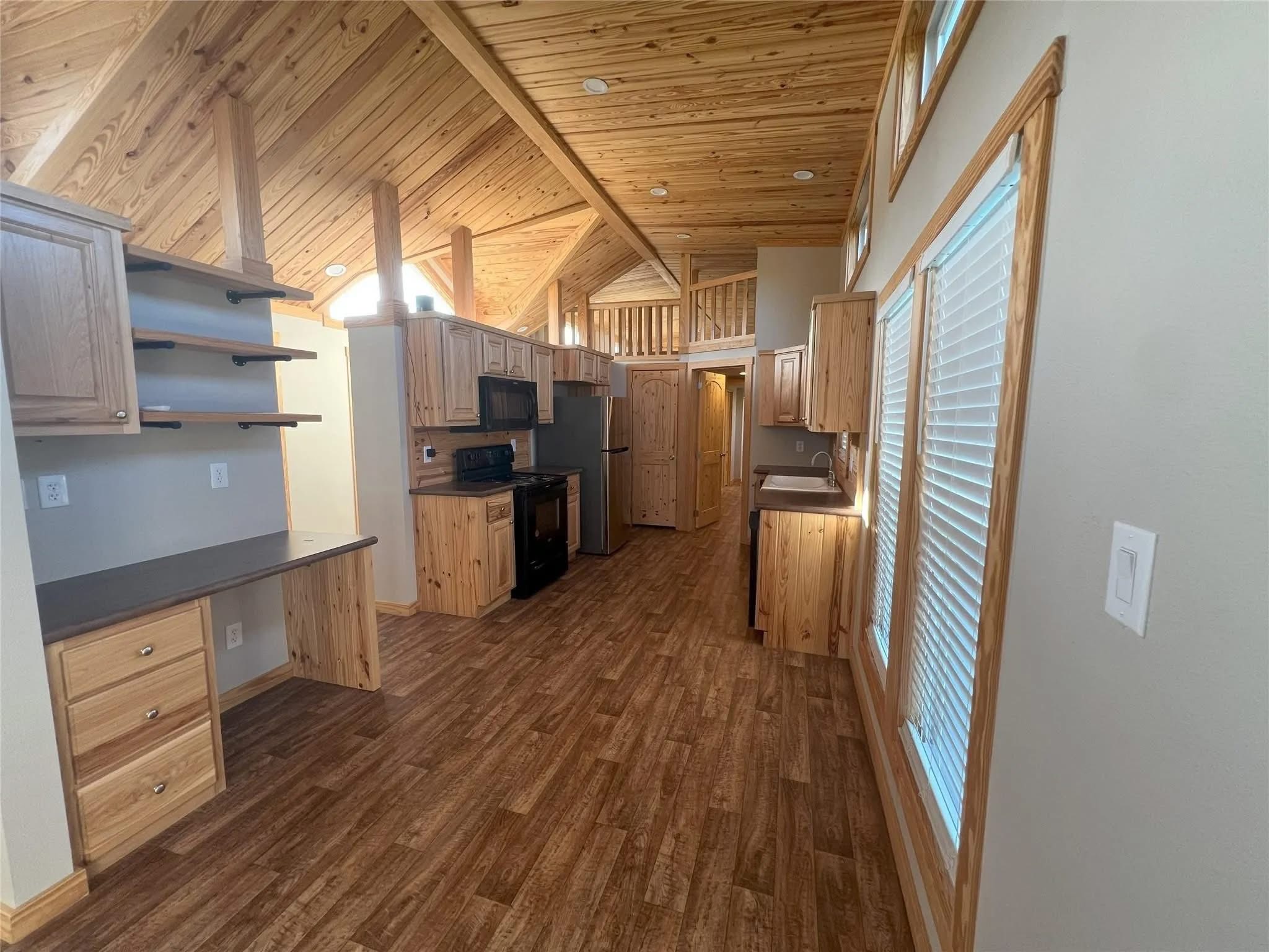 Warm wooden kitchen interior with high vaulted ceilings. Features include light wood cabinets, black appliances, and large windows creating an inviting, airy space.