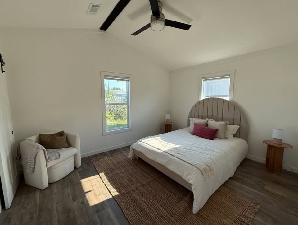 Cozy bedroom with a modern aesthetic. Features a beige bed with a rounded headboard, warm pillows, armchair, woven rug, wood side tables, ceiling fan, soft daylight.