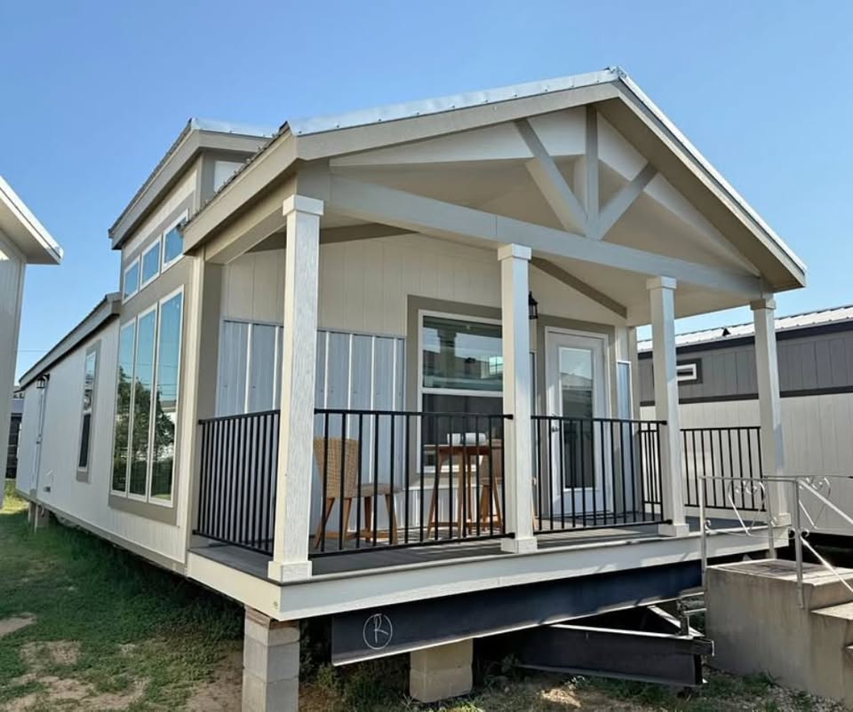 Small modern tiny house with a covered porch, black railing, and tall windows. Bright, sunny day, and two wicker chairs on the porch suggest relaxation.