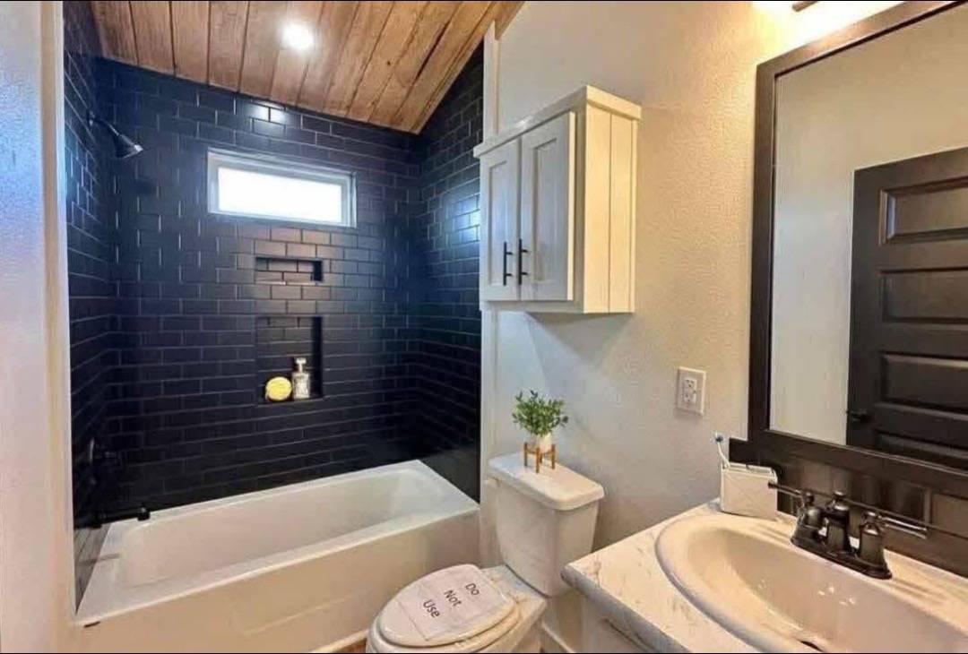 Cozy bathroom with wood ceiling, black tile wall, white tub. Vanity features black-framed mirror, potted plant, and a white cabinet. Modern and sleek.