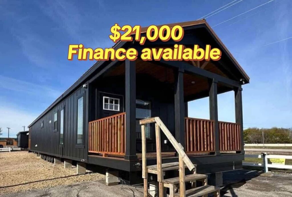 A small, black wooden cabin with a front porch and stairs is displayed under a bright blue sky. Text overlay: "$21,000 Finance available."