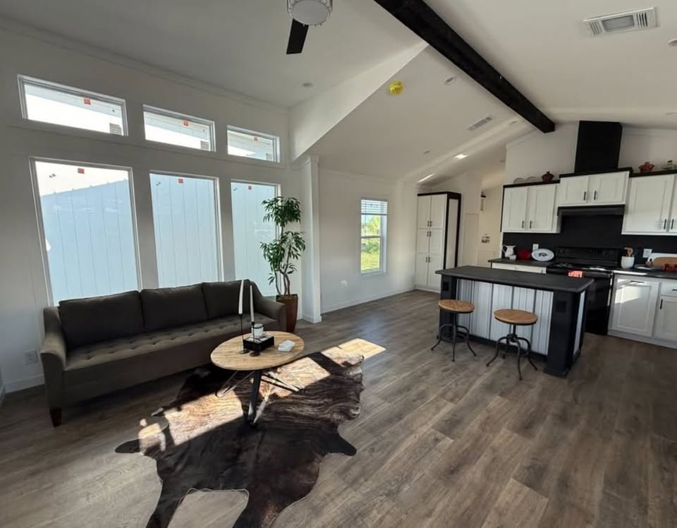 Modern open-plan living area with wooden flooring, featuring large windows, a gray sofa, round table with cowhide rug, and a kitchen with a black island.