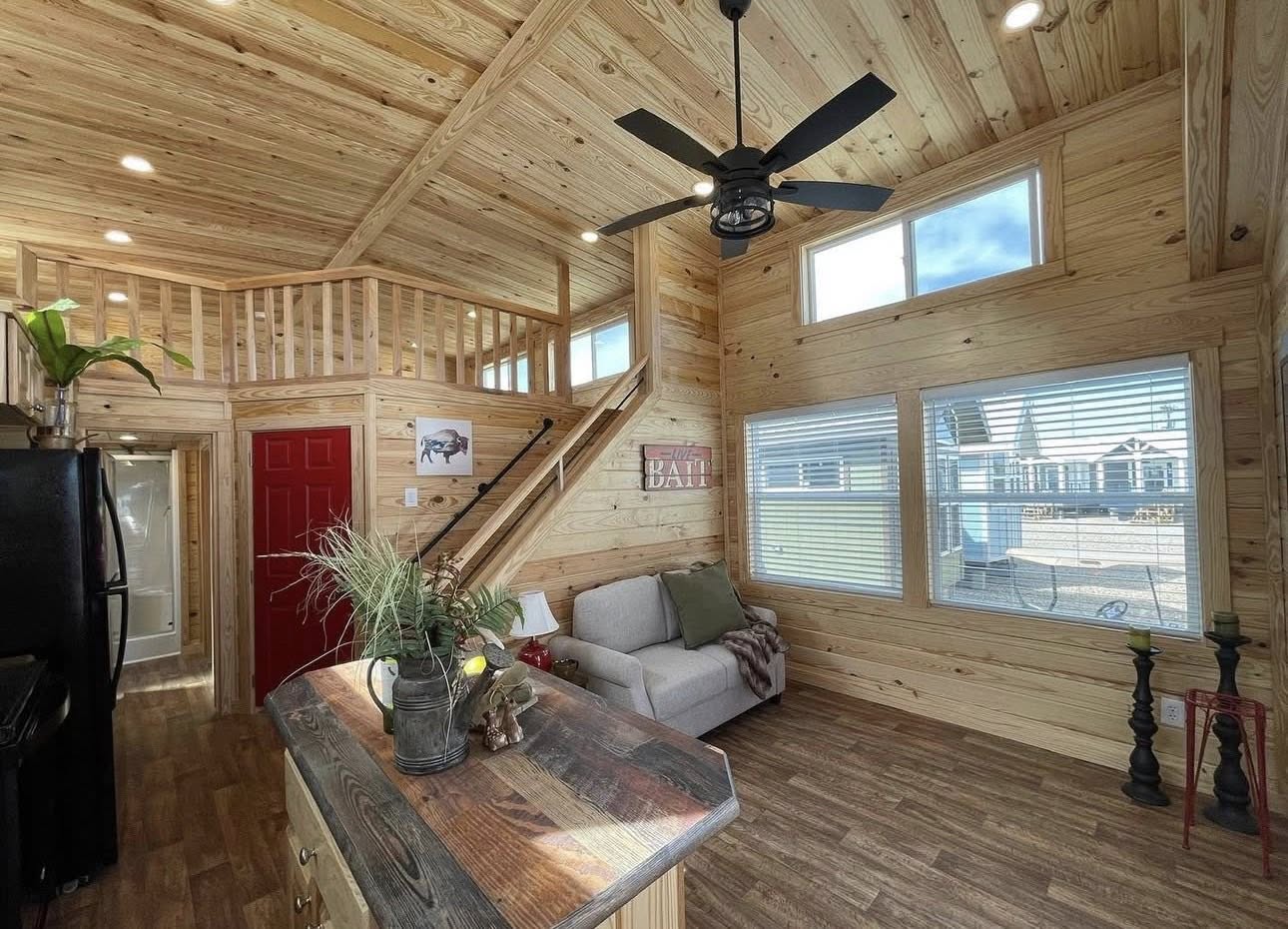 Cozy wooden tiny home interior with loft, red door, and large windows. Features a sofa, ceiling fan, and rustic decor on a wooden island. Bright and inviting.