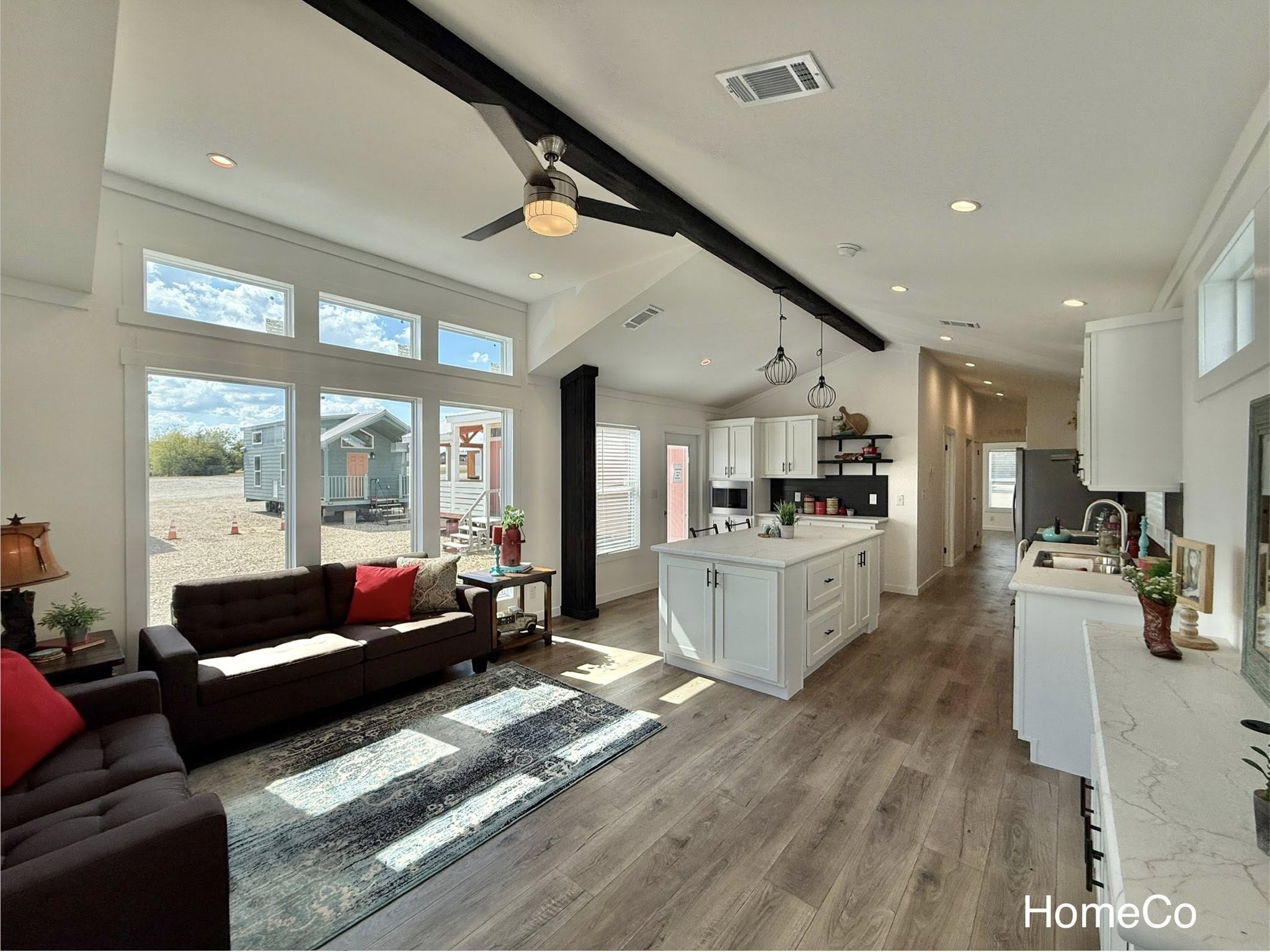 Spacious, airy living area with modern kitchen, large windows, and rustic beams. Cozy sofas with red cushions create a warm, inviting atmosphere.