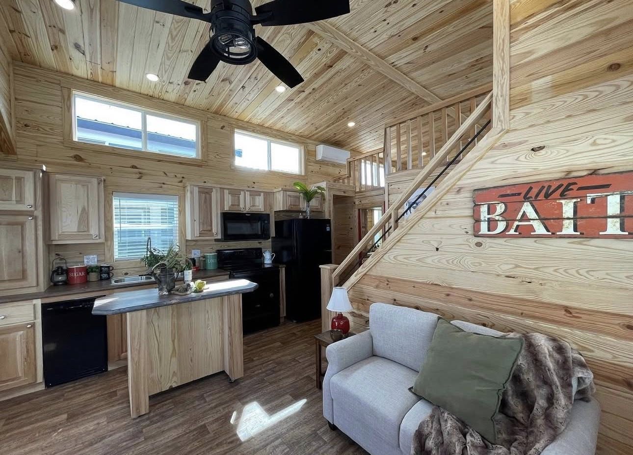 Bright tiny home interior with wooden walls and ceiling. Cozy kitchen and living area with a sofa, island, and staircase. Text on wall reads "Live Battle."