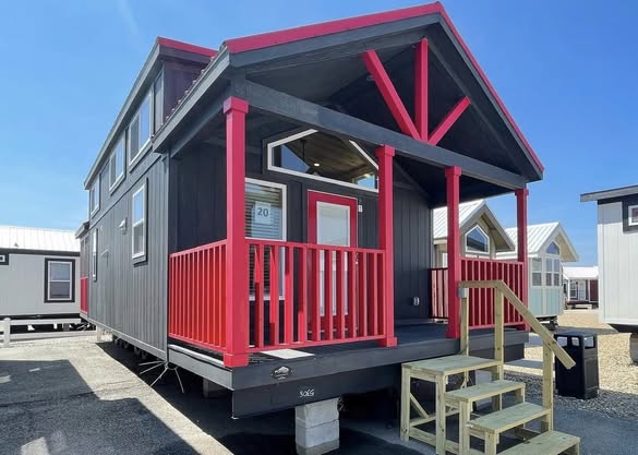 A modern tiny house with a dark exterior accented by vibrant red trim and porch railing. Wooden steps lead to the entrance, set under a clear blue sky.