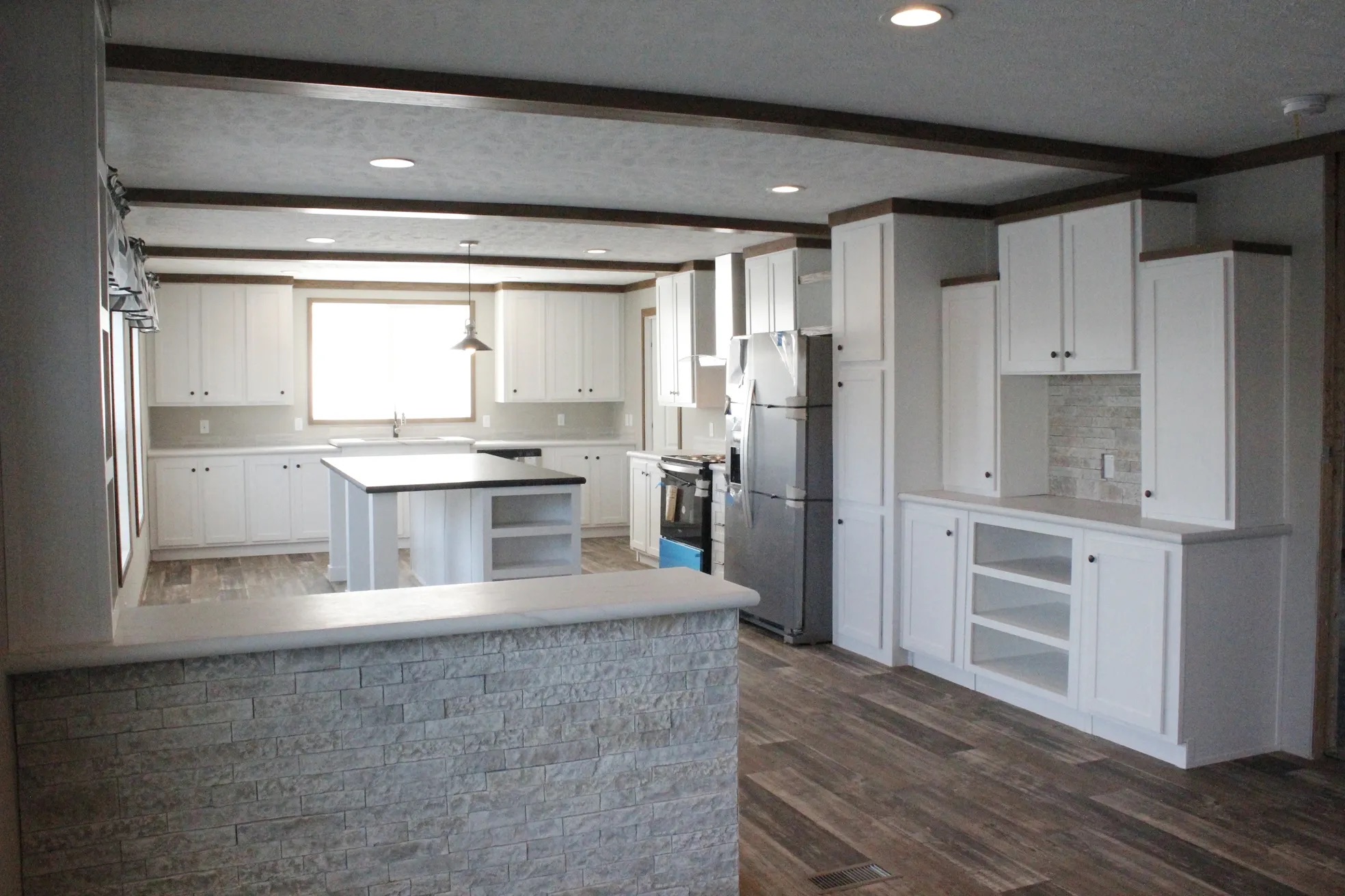 Spacious kitchen with white cabinets, stone accent wall, stainless steel appliances, and wooden flooring. Central island under hanging light, bright and modern.