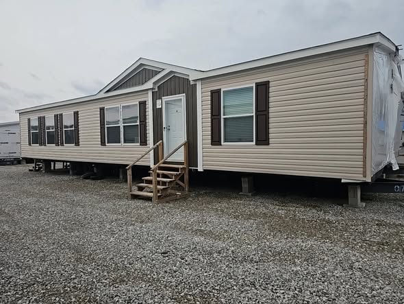 A beige manufactured home with brown shutters sits on a gravel lot. It has a white front door and wooden steps, conveying a practical, rustic tone.