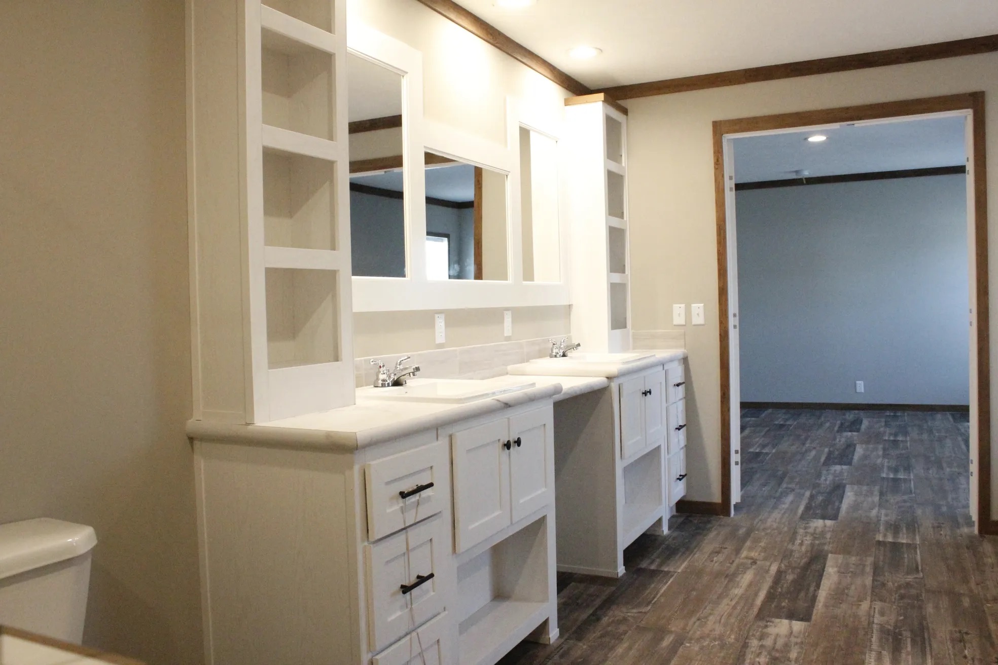 Spacious bathroom with dual sinks, white cabinets, open shelving, and a large mirror. Light gray walls and wood-textured flooring create a modern feel.
