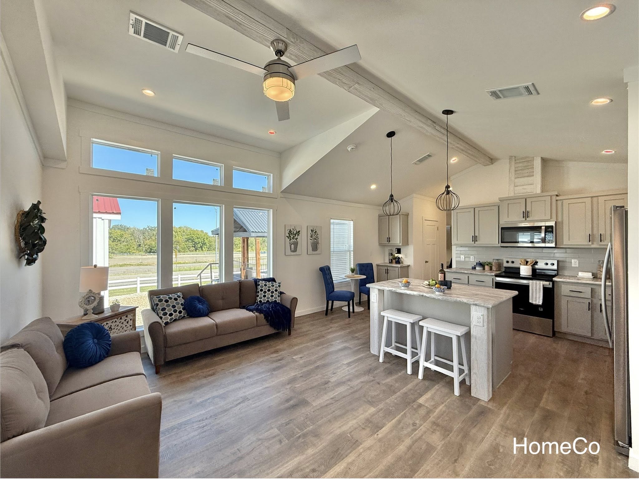 Spacious open-concept modern coastal farmhouse interior featuring vaulted beamed ceiling with ceiling fan, large floor-to-ceiling windows flooding natural light, bright white shiplap walls, light gray luxury vinyl plank flooring, neutral beige sofa with blue accent pillows, quartz-top kitchen island with white shaker cabinets, gray subway tile backsplash, stainless steel appliances, pendant lighting, cozy living-kitchen-dining flow, blue upholstered chairs, potted plants, and scenic outdoor views creating an airy bright family-friendly living space.