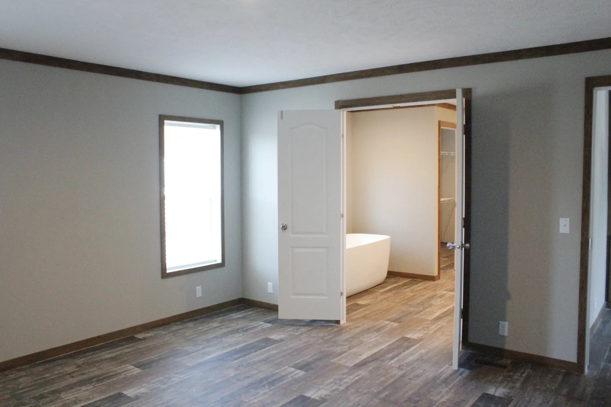 A minimalistic room with light gray walls and wooden flooring. A window lets in natural light. An open door reveals a modern bathroom with a bathtub.