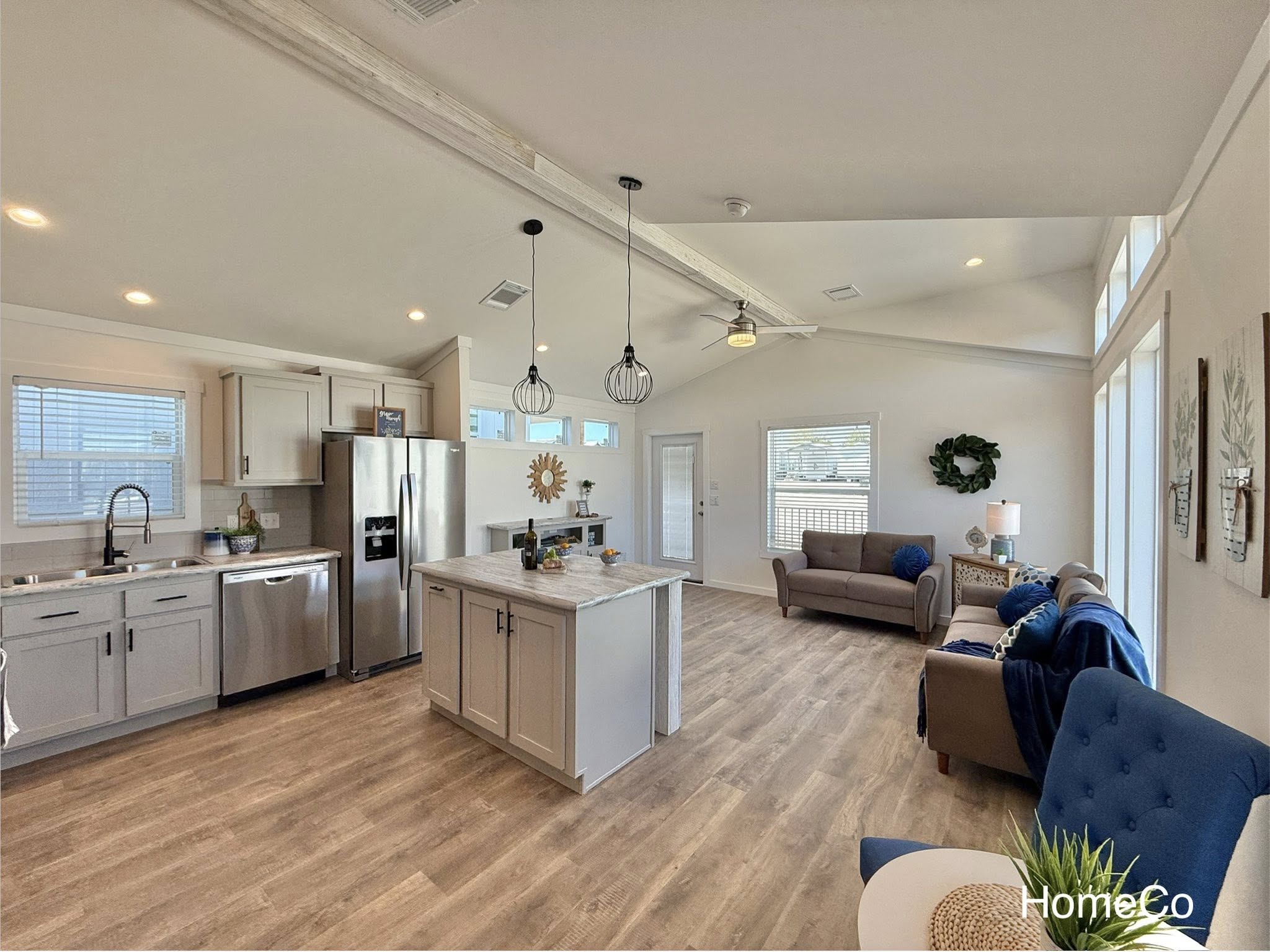 Spacious bright modern farmhouse open-concept kitchen living room combo with vaulted ceiling featuring exposed rustic wooden beams, ceiling fan and pendant lights, light gray shaker cabinets, large quartz waterfall island with seating, stainless steel appliances including side-by-side refrigerator, farmhouse sink with black faucet, gray subway tile backsplash, luxury vinyl plank flooring in warm neutral tones, cozy sectional sofa in beige with blue throw pillows and blanket, accent blue tufted chair, wooden sunburst decor, potted plants, fresh fruit bowl, high clerestory windows and large sliding doors flooding natural light, airy inviting family gathering space perfect for coastal cottage or tiny home living.