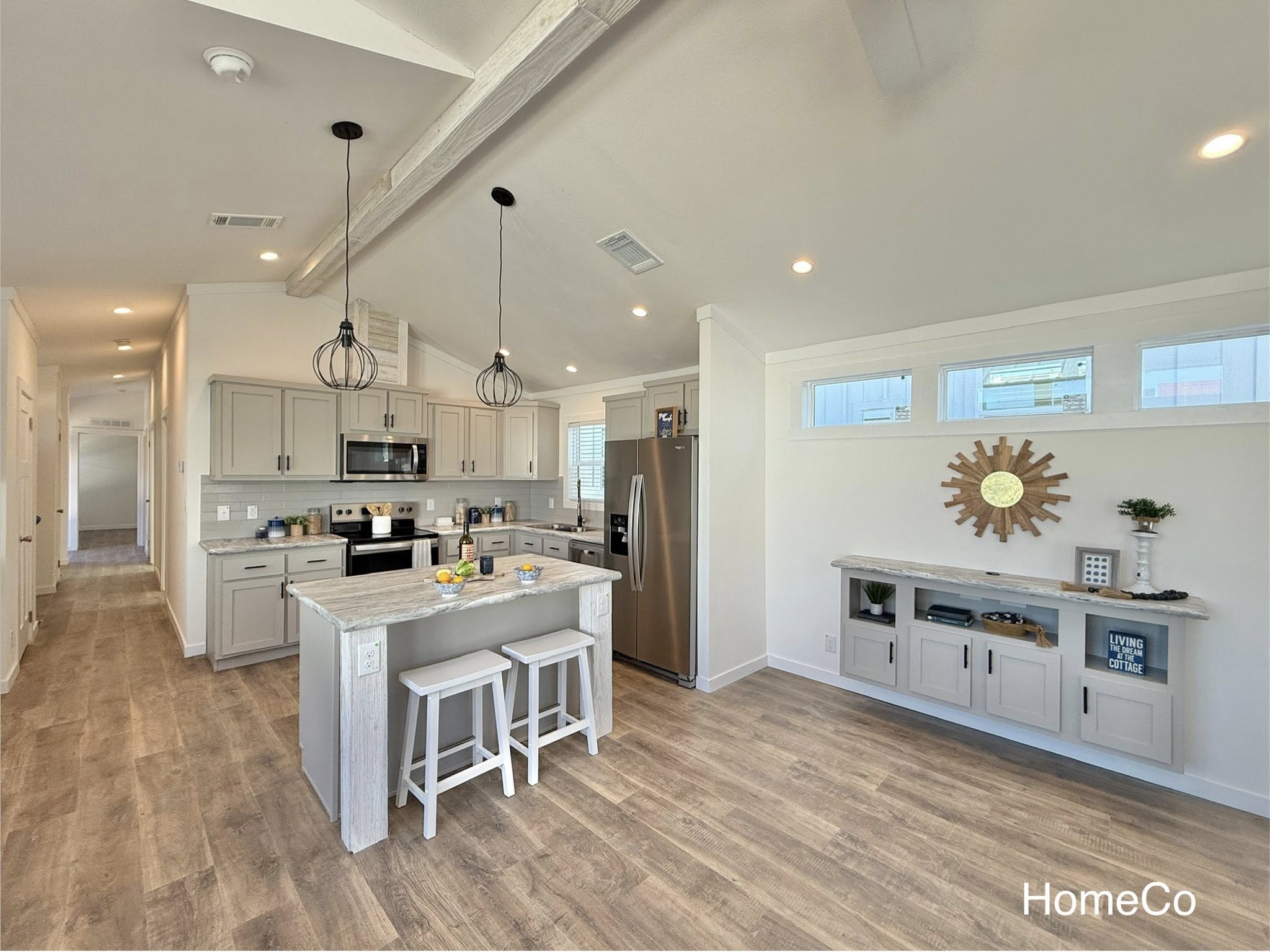 Bright and airy modern farmhouse open-concept kitchen and living space featuring vaulted ceiling with exposed rustic wooden beams, pendant lighting over large gray quartz island with white shaker base cabinets, stainless steel appliances including double-door refrigerator and gas range, gray subway tile backsplash, light gray luxury vinyl plank flooring, neutral beige walls, hallway entry flow, built-in console with marble top and open shelving displaying "Living the Dream at the Cottage" sign, wooden sunburst wall decor, potted plants, fresh fruit bowl, high clerestory windows for abundant natural light, spacious layout ideal for family gatherings and cozy cottage-style living.