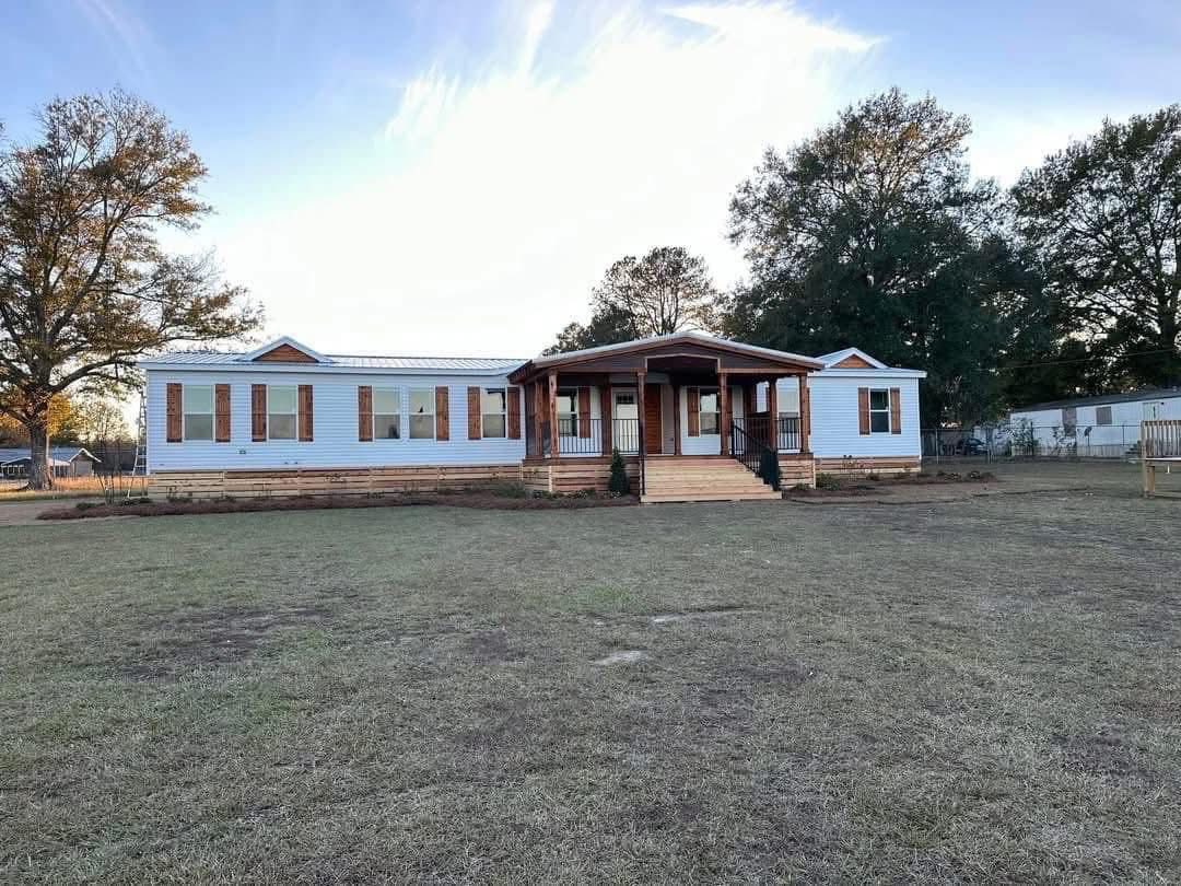 Spacious white manufactured home with a wooden porch sits amid a large grassy yard. Tall trees frame the scene under a clear, blue sky.