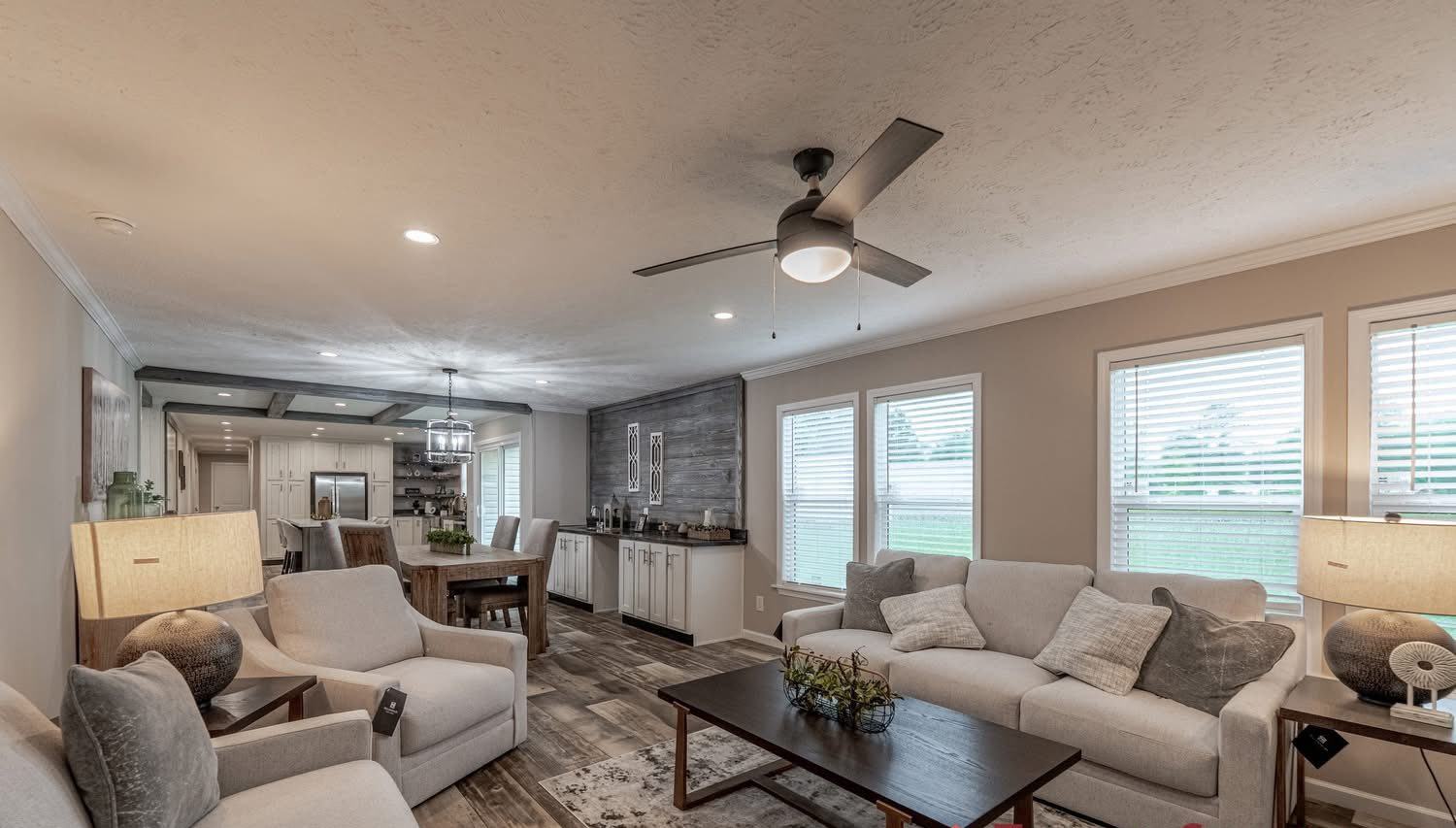 Modern living room with neutral tones, featuring gray sofas, a black coffee table, and ceiling fan. Large windows bring in natural light. Cozy and inviting.