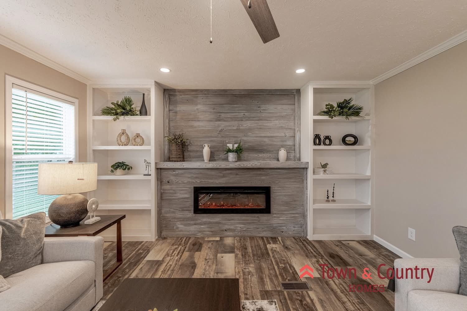 Cozy living room with a modern electric fireplace and wood paneling. White shelves hold plants and decor. Neutral tones create a calm ambiance.