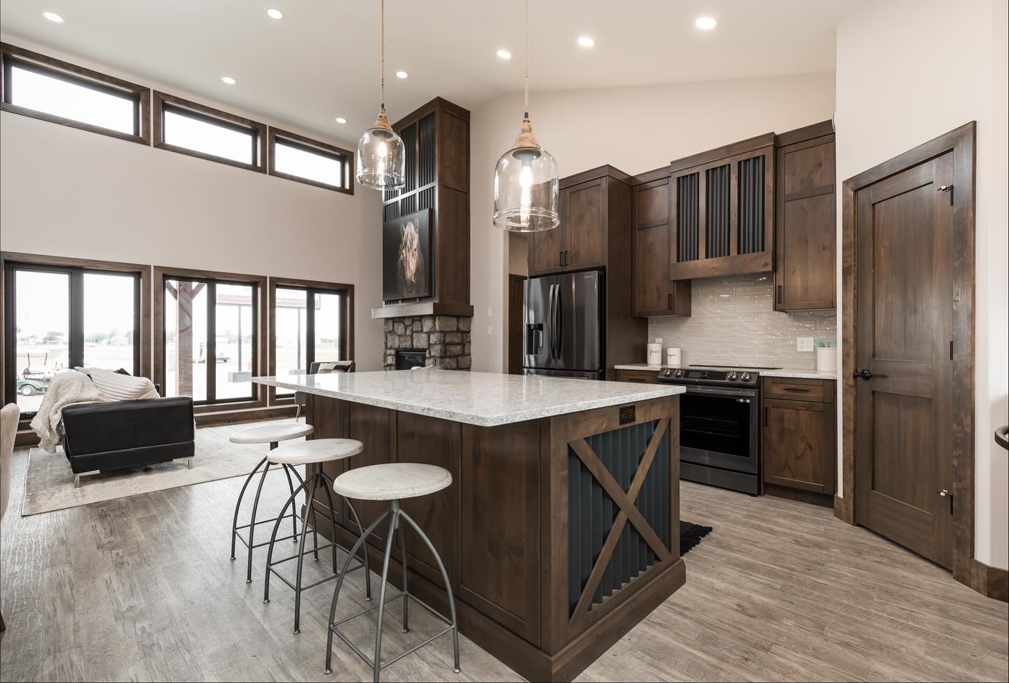 Modern kitchen with rustic charm, featuring wooden cabinets, stone fireplace, marble island with barstools, and large windows, creating a cozy atmosphere.