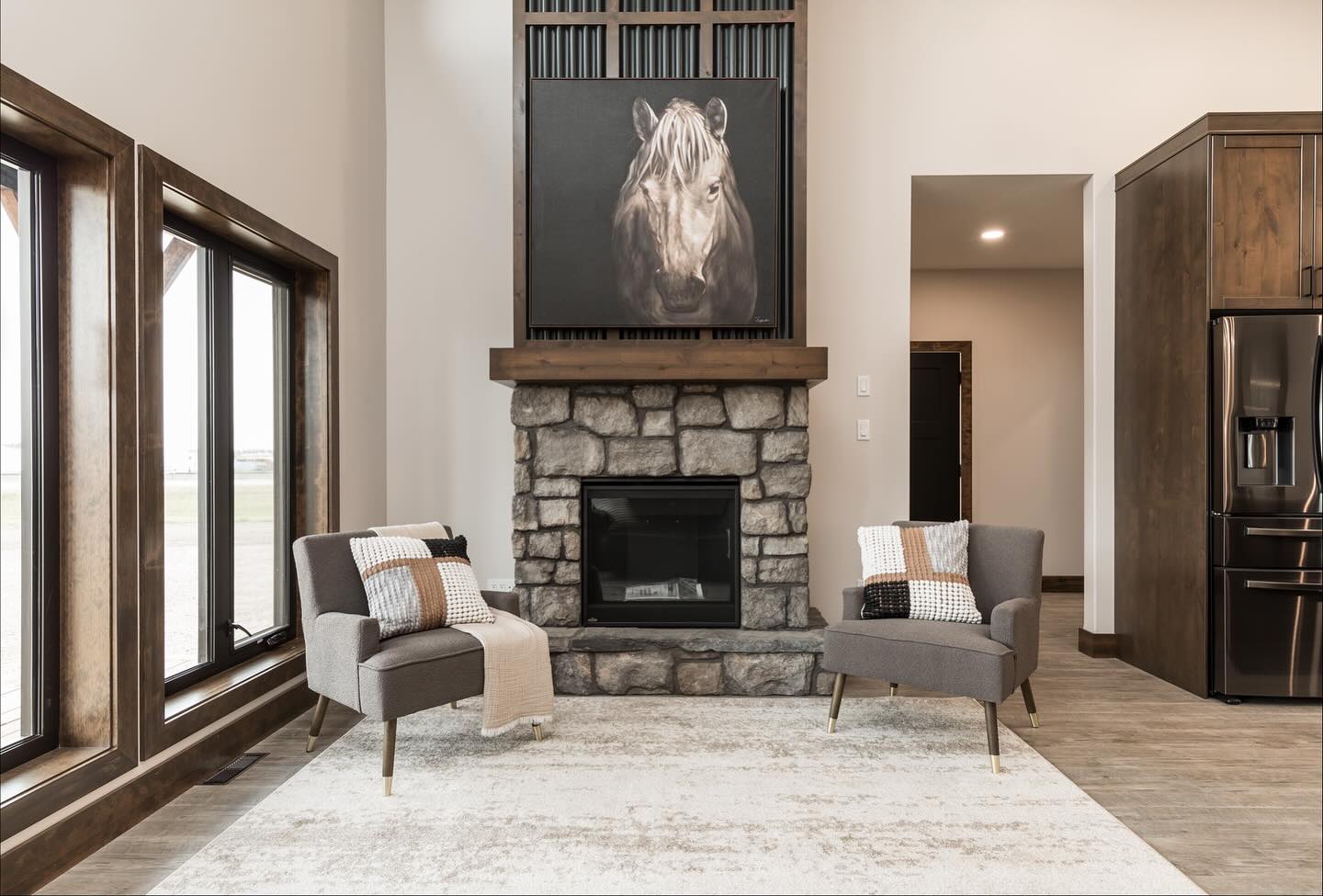Modern living room with stone fireplace, horse painting above. Gray chairs with blankets face each other on a light rug. Large windows on the left.