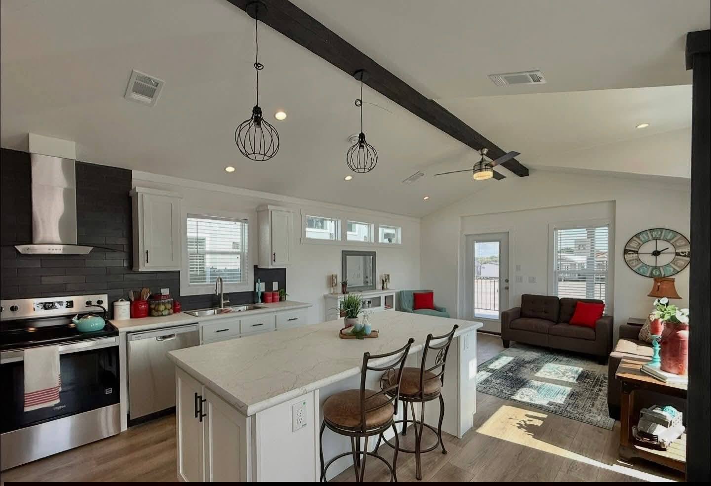 Modern open kitchen-living area with a large island and two barstools. Black accents, high ceilings, and natural light convey a cozy, stylish atmosphere.