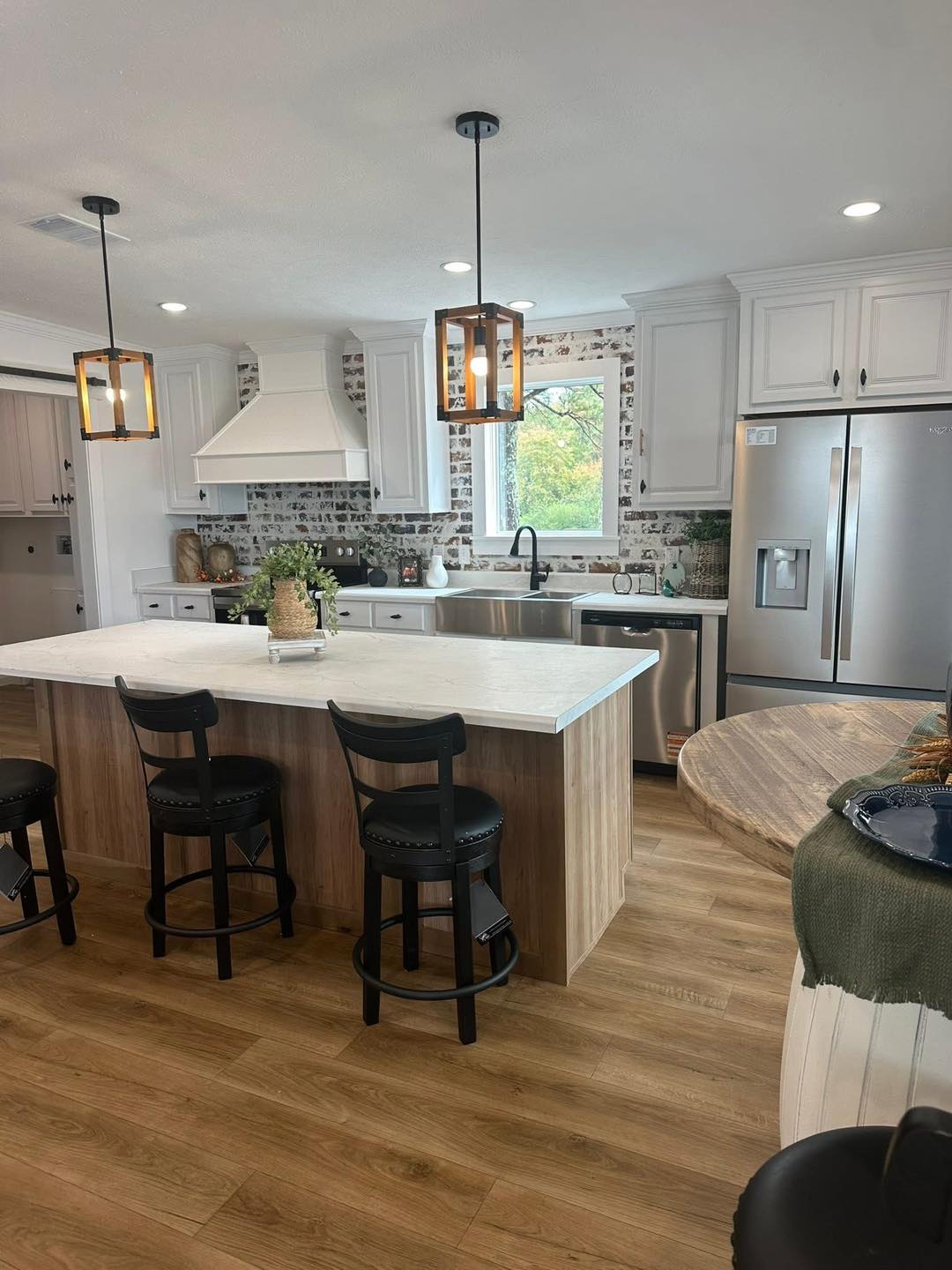 Modern kitchen with wooden flooring features a large island, three black chairs, stainless steel appliances, white cabinets, and pendant lights.