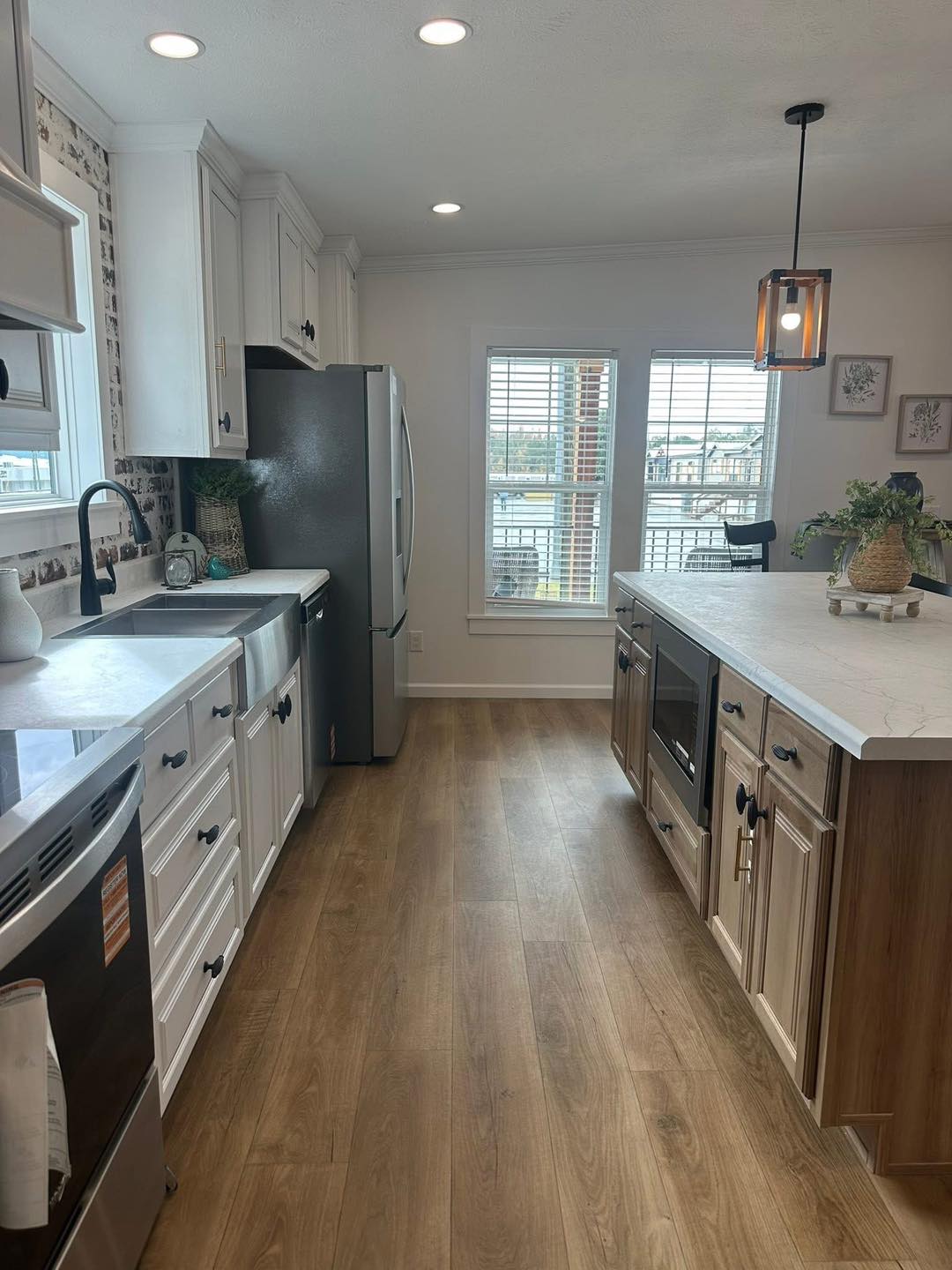 Bright kitchen with white cabinets, stainless steel appliances, and wood flooring. A center island features a pendant light above. Large windows bring in natural light. Elegant, cozy, and inviting atmosphere.