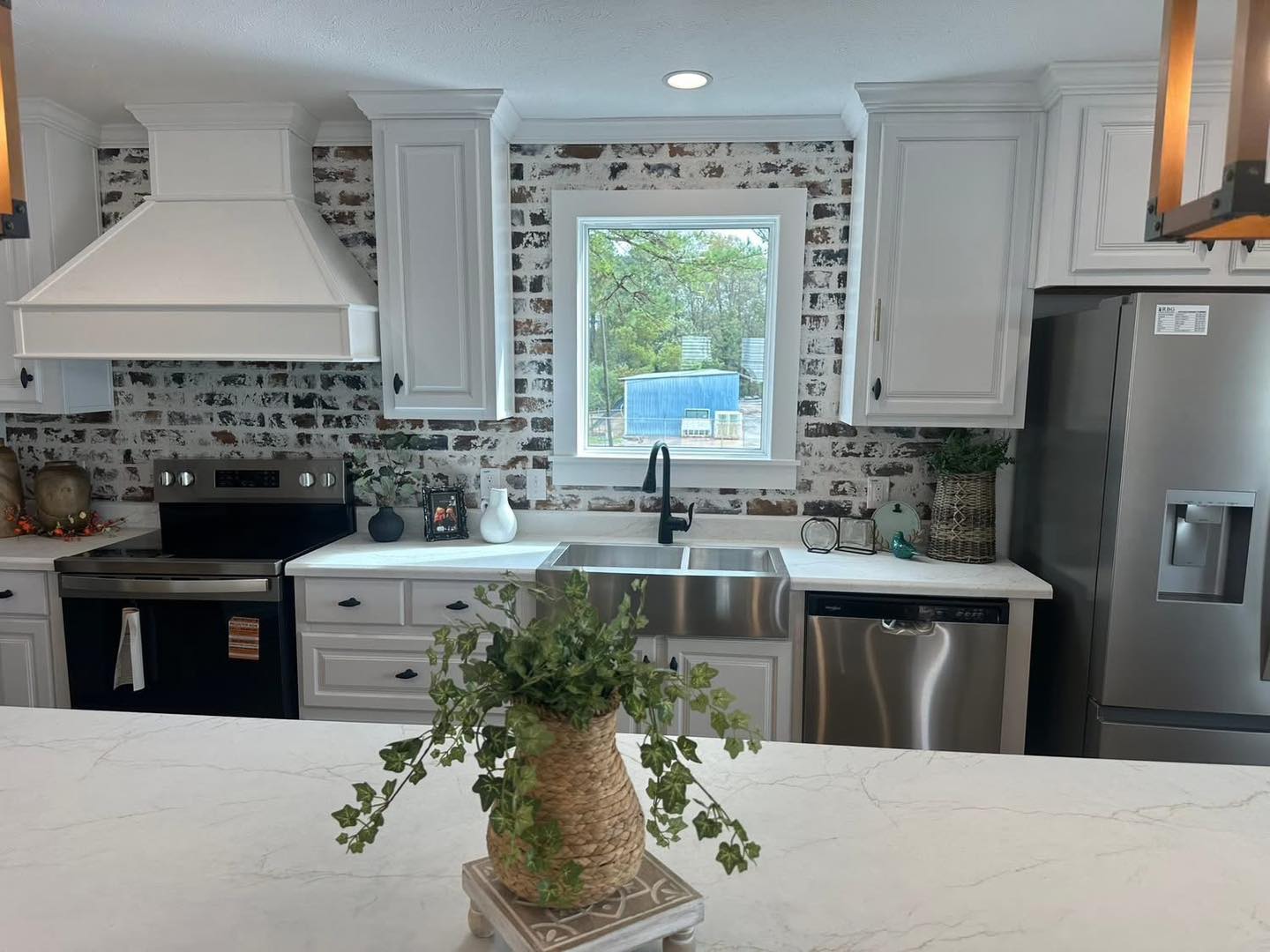 Bright kitchen with a white range hood, stainless steel appliances, and rustic brick backsplash. A window with a garden view centers the room.