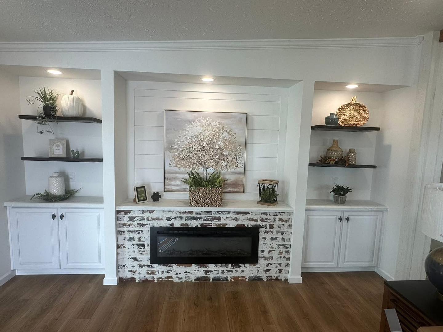 Cozy living room with a white brick fireplace, framed tree artwork, and built-in shelves holding decorative items like plants and baskets. Warm, inviting tone.