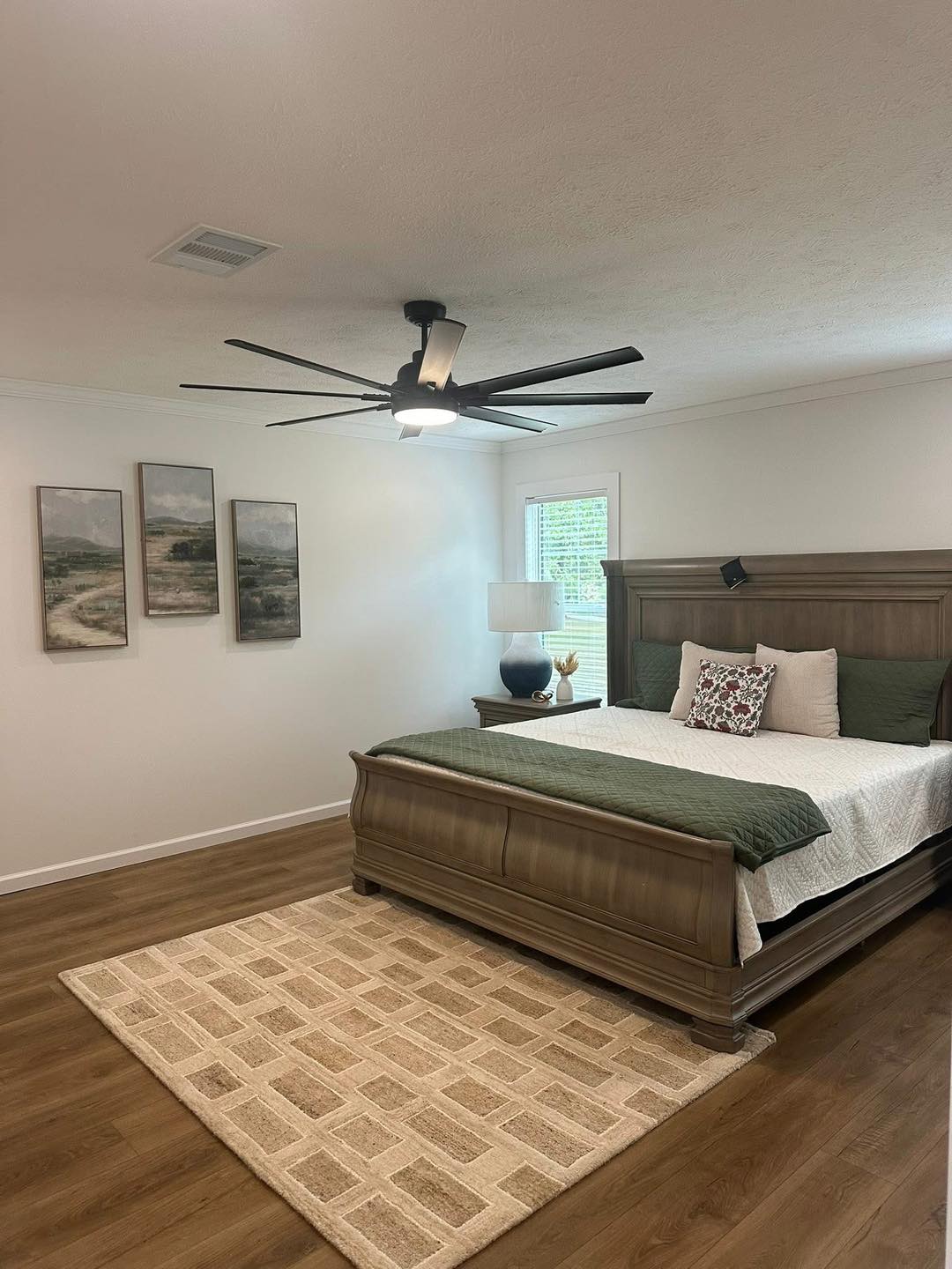Spacious bedroom with a large wooden bed, green and white bedding, and decorative pillows. A ceiling fan and a lamp add warmth. Art on the white wall.