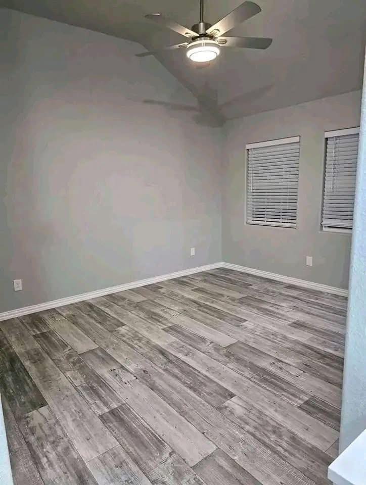 Empty room with gray walls and wood-look tile flooring. Two windows have closed blinds, and a ceiling fan with a light fixture hangs above.