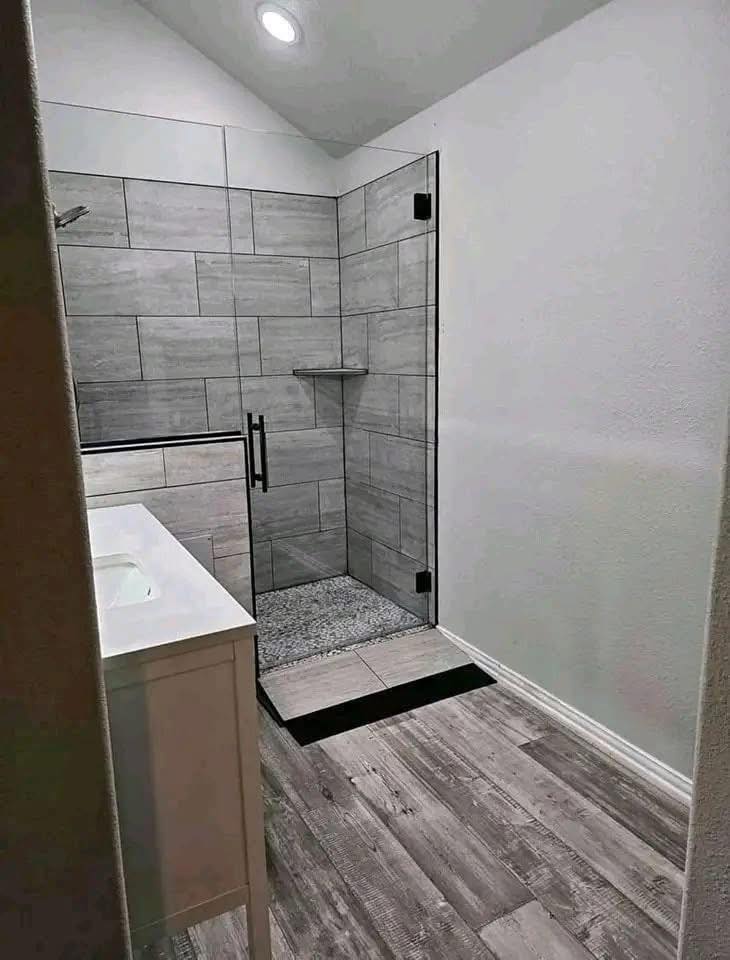 Modern bathroom featuring a spacious walk-in shower with gray tiles and a glass door. The floor is wood-style, and a white sink counter is visible. Minimalist and clean.