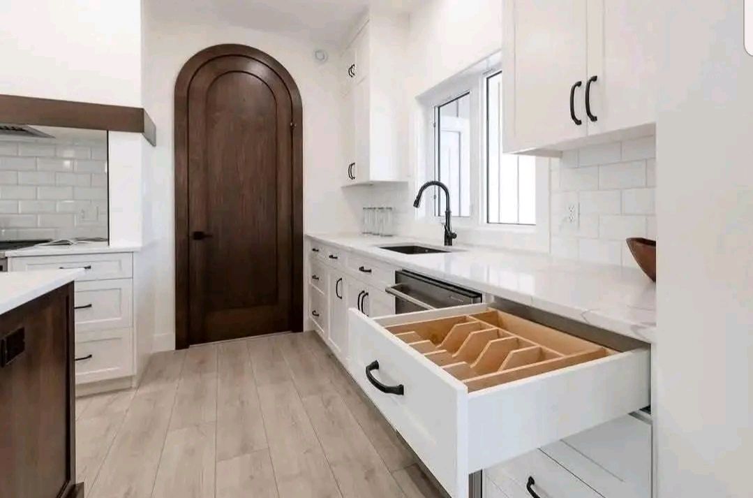A modern kitchen with white cabinets, dark handles, and a wooden floor. An open drawer reveals organized dividers. A dark arched door adds contrast.
