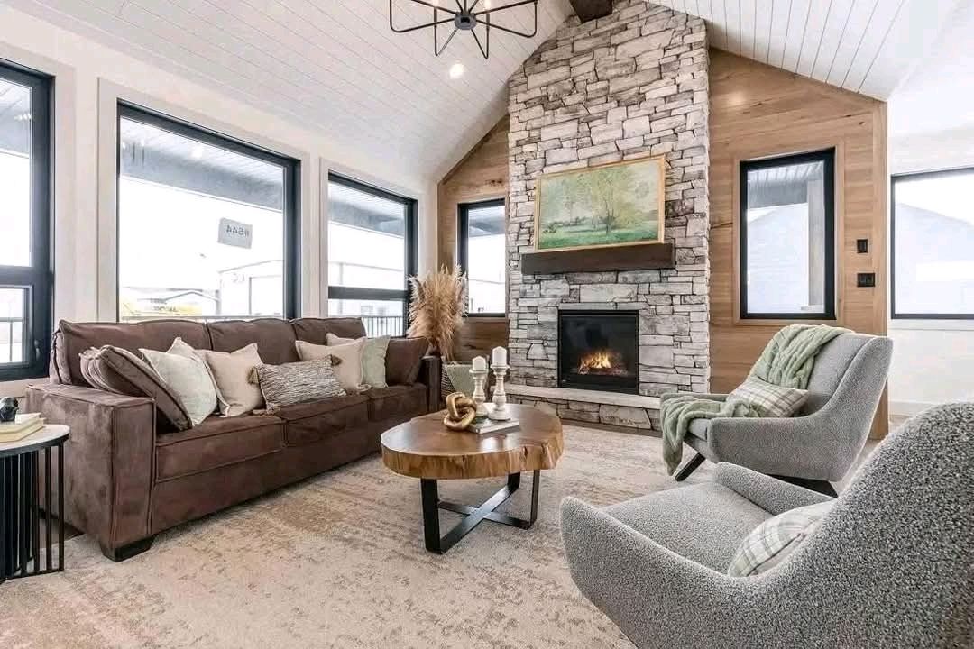 Modern living room with a cozy atmosphere. Brown couch with pillows, stone fireplace, abstract art, round wooden coffee table, and gray armchairs.