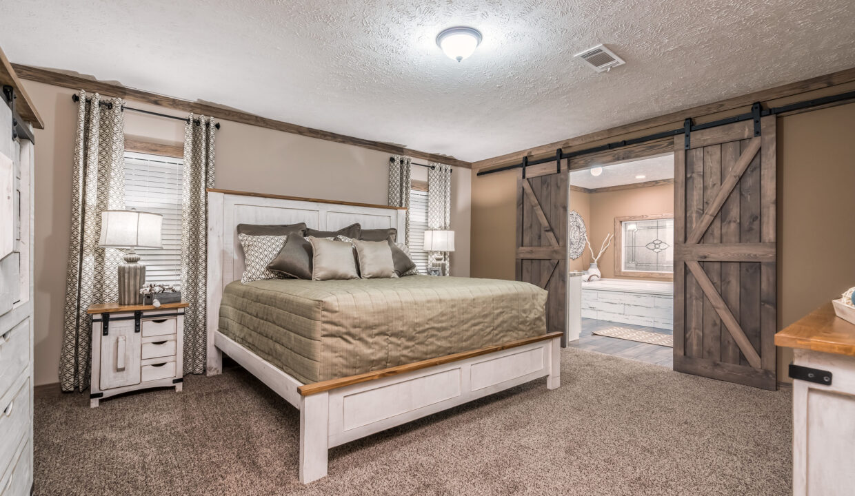 Spacious bedroom with a rustic feel, featuring a large bed with beige bedding, wooden barn doors leading to a bathroom, and warm lighting from bedside lamps.