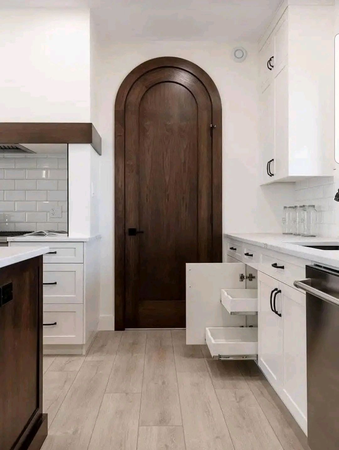 Sleek kitchen with white cabinets, black handles, and a high arched wooden door. Open drawers reveal a clean, minimalist design. Light wood floors add warmth.